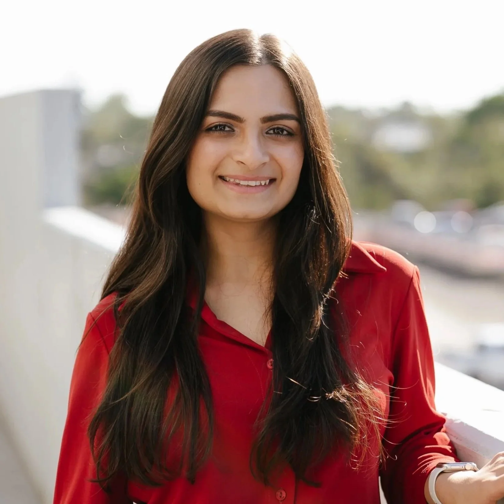 A woman with long dark hair, wearing a red top, is smiling outdoors near a white railing with a blurred cityscape background.