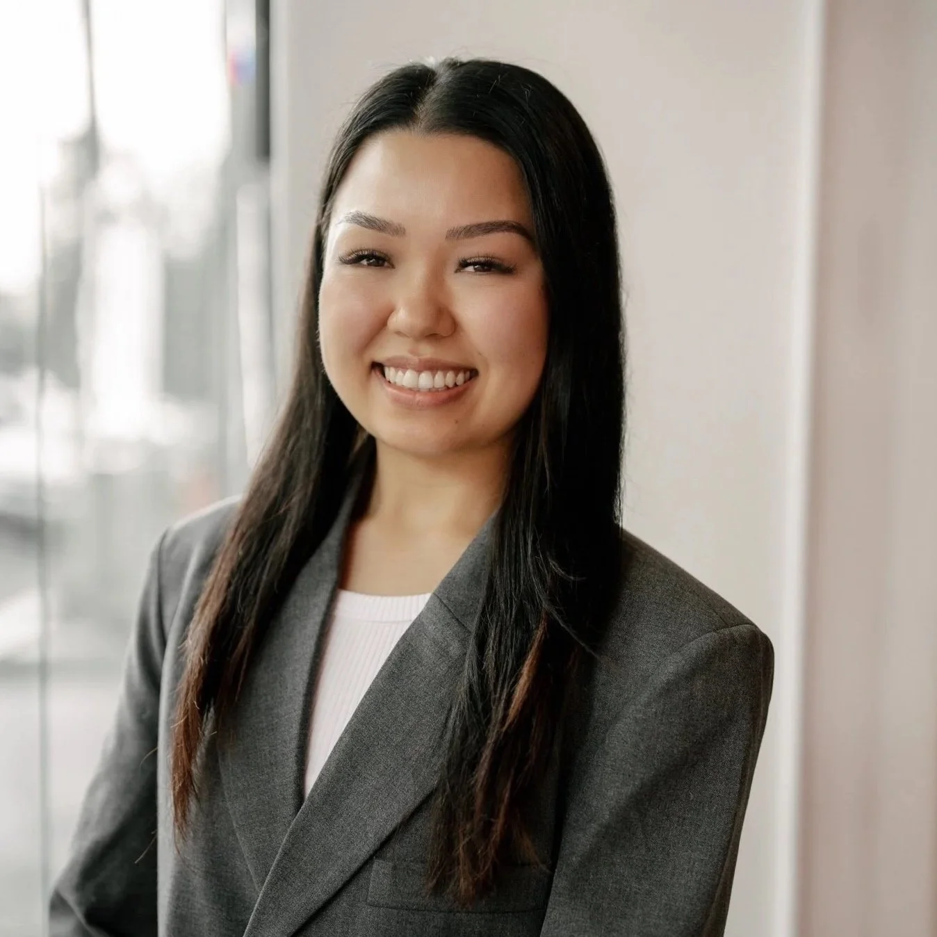 A young woman with long dark hair, smiling, wearing a gray blazer and white top, standing indoors near a window.