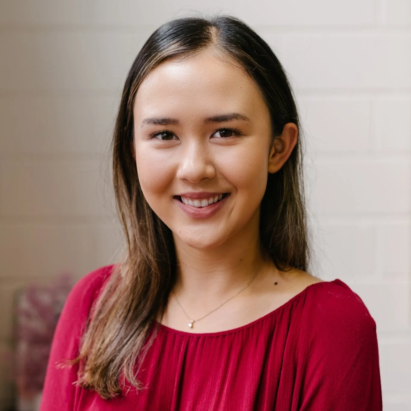 A young woman with long brown hair and a cheerful smile, wearing a red top and a delicate necklace, standing in front of a light-colored, horizontal paneled wall.