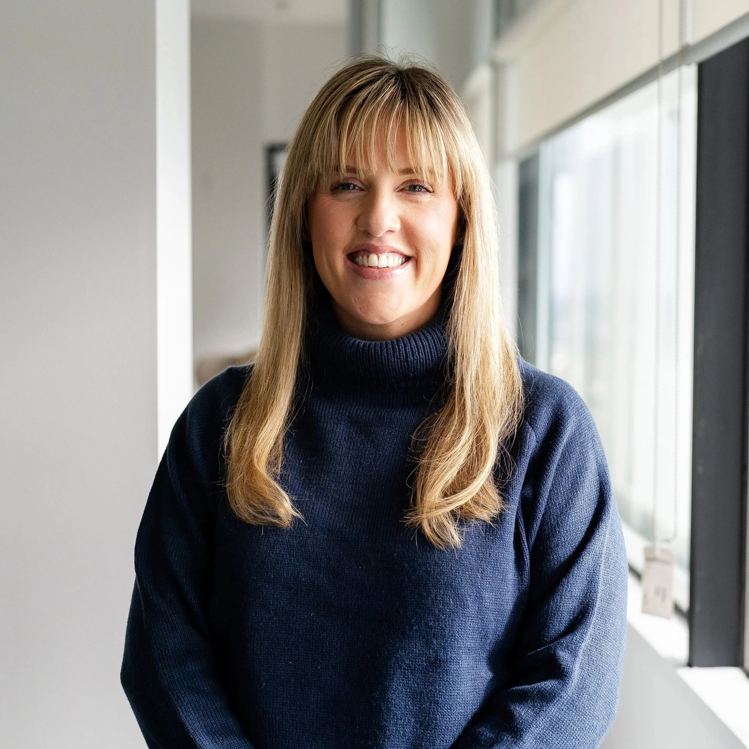 Close up of woman with long blonde hair and a straight cut fridge, wearing a navy turtleneck knitted jumper. She stands in front of a window with natural light.