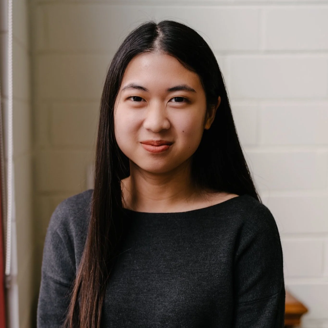 A young woman with long black hair, wearing a dark gray top, standing in front of a white brick wall.