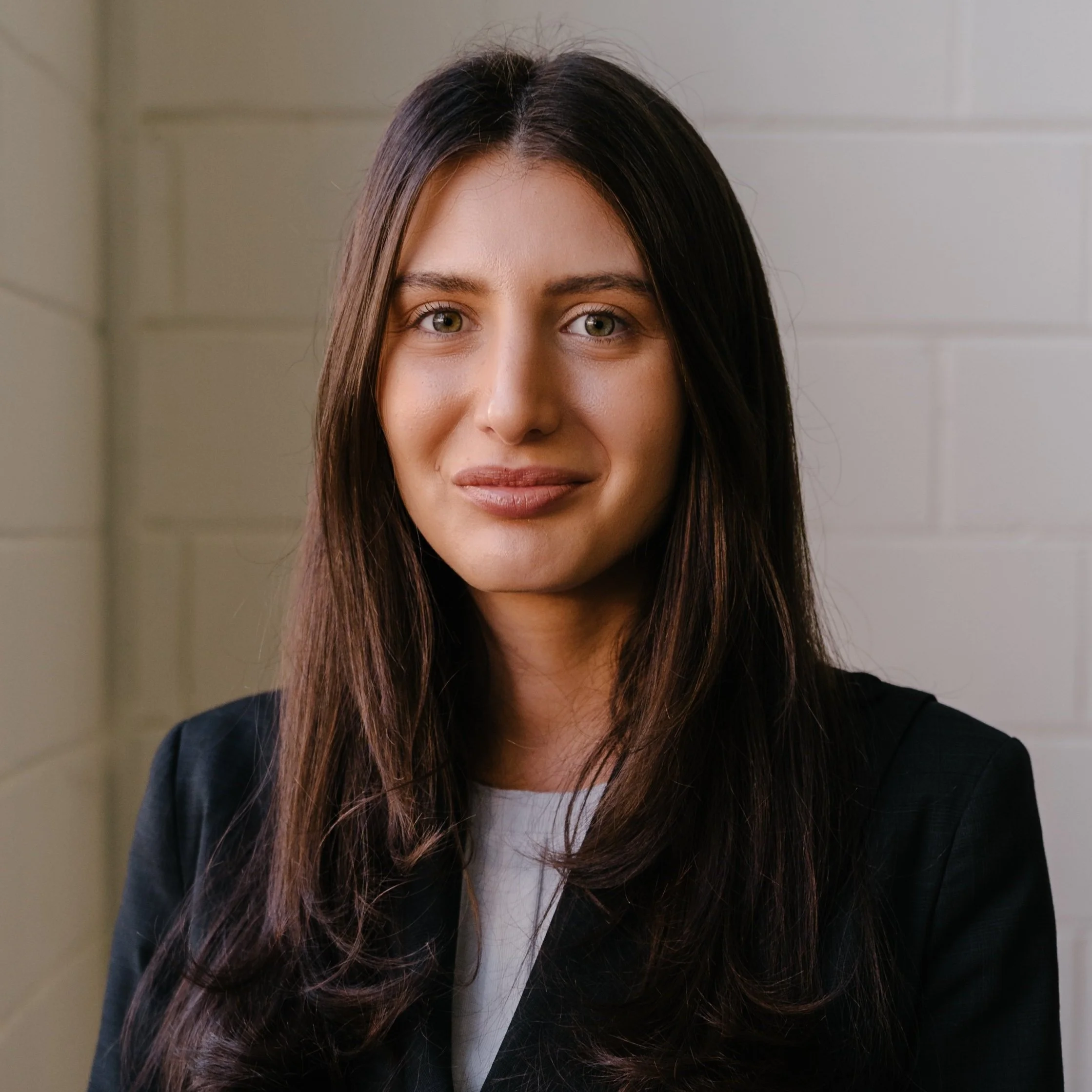 Close-up of a young woman with long dark hair and hazel eyes, smiling softly, wearing a black blazer and white top, standing against a plain background.