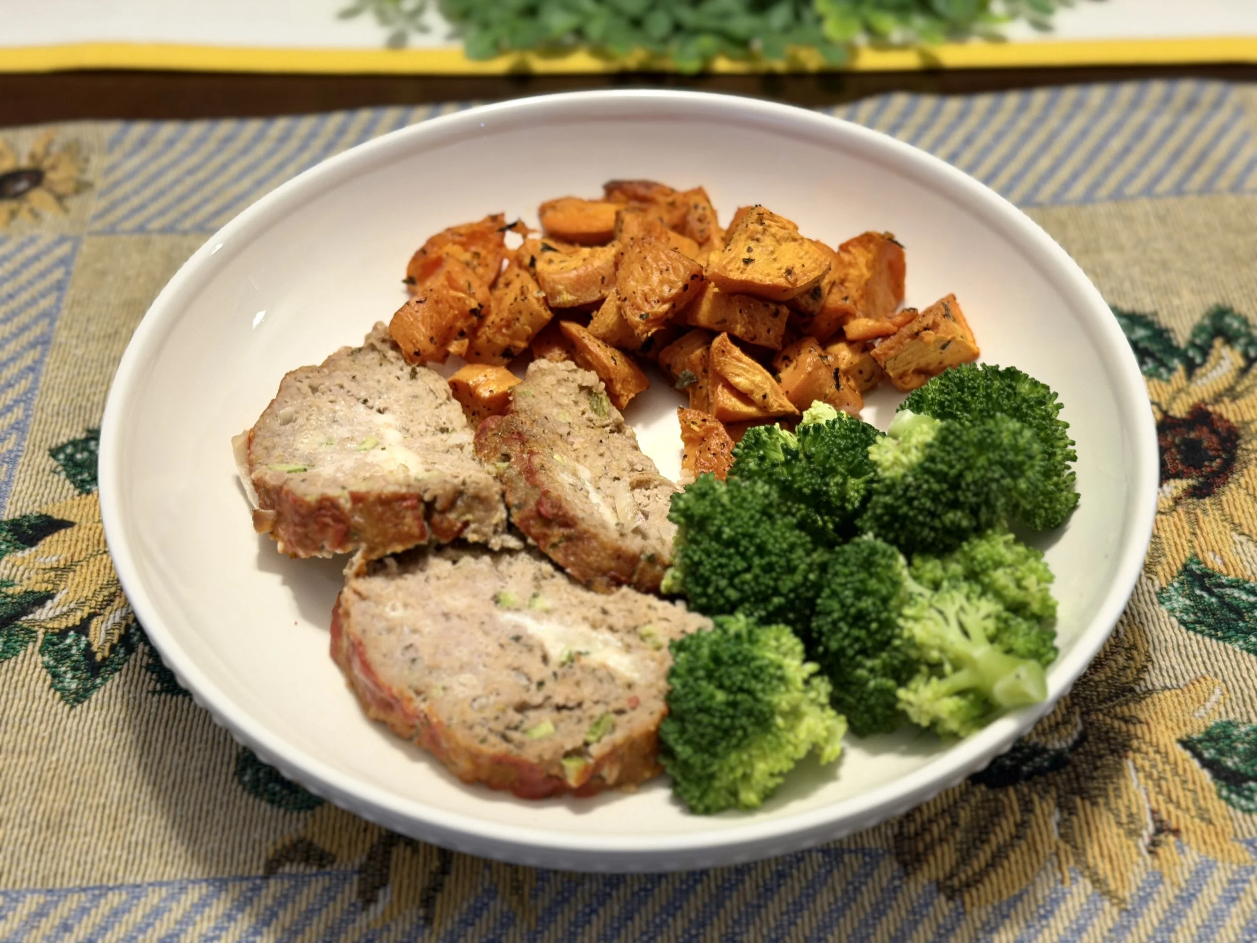 A white bowl containing cooked sweet potatoes, meatloaf slices, and fresh green broccoli on a patterned tablecloth.