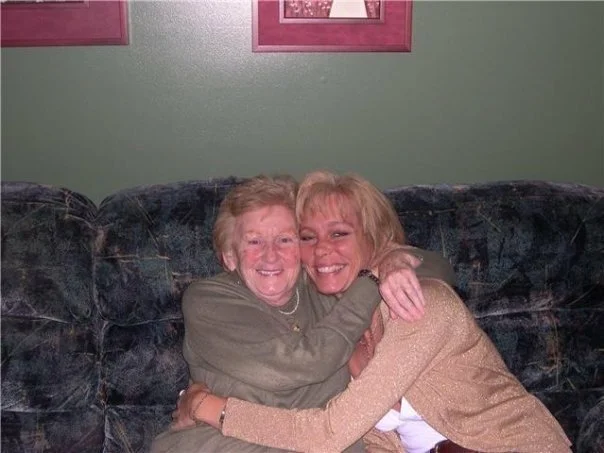 Two women hugging and smiling on a sofa inside a room with green walls and pink picture frames.