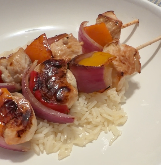 Skewers of grilled chicken, red onion, red bell pepper, yellow bell pepper, served over white rice.
