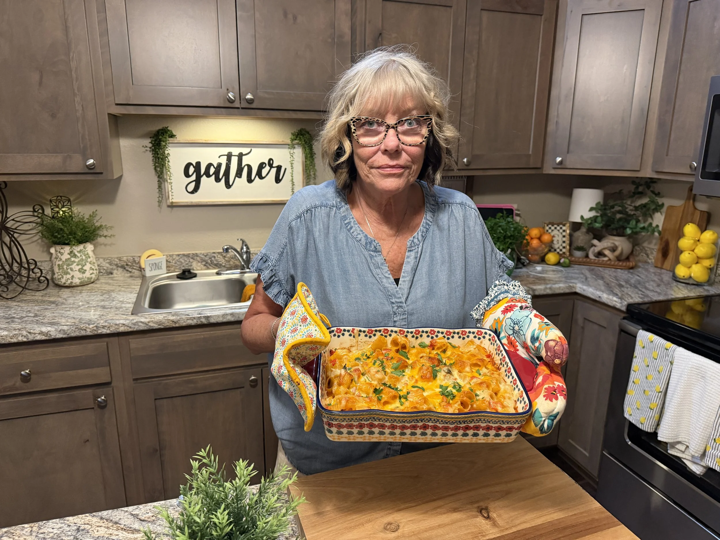 An older woman with blonde and dark brown hair, wearing leopard print glasses and a blue denim shirt, holding a colorful baking dish with baked pasta topped with cheese and herbs in a kitchen.