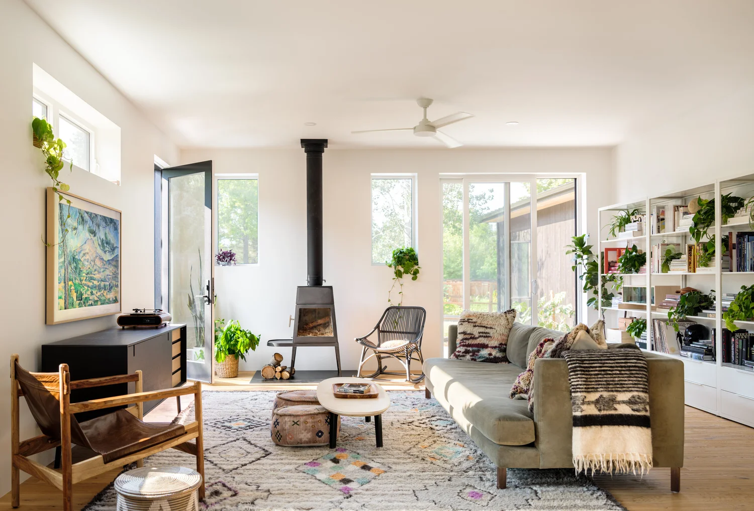 Living room with pale upholstered seating, black stove, layered rugs, wood flooring, and leafy plants.