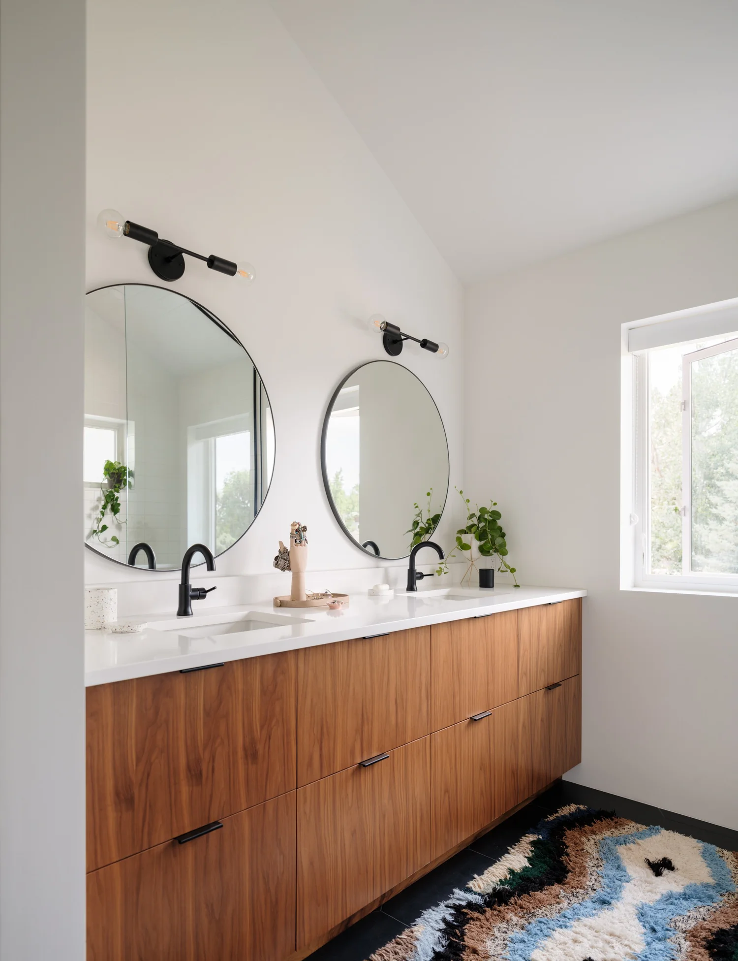 Bathroom with a long wood vanity, white counter, twin round mirrors, black wall lights, and plants.