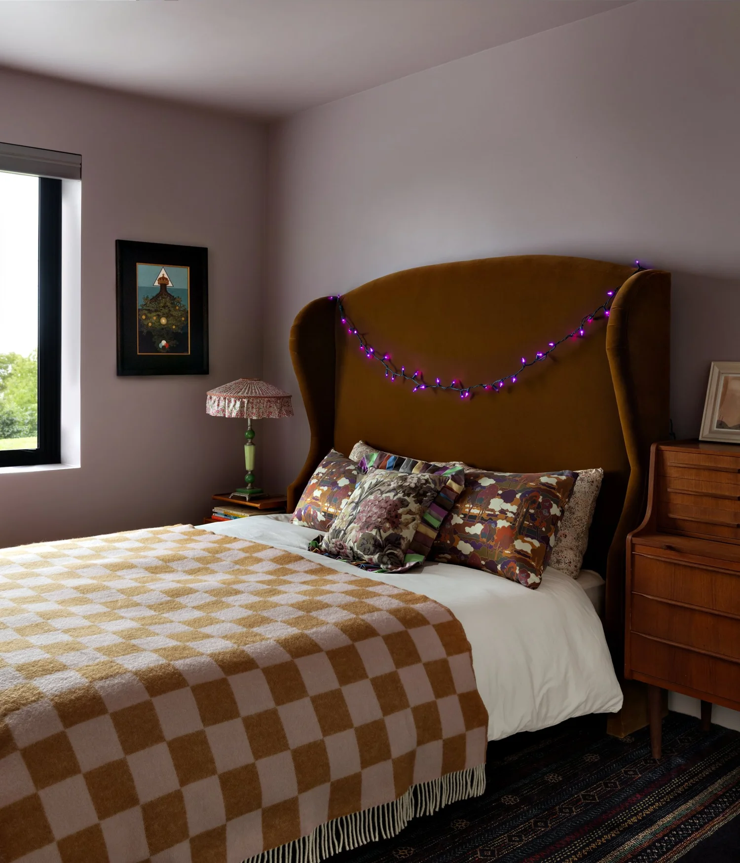 Bedroom with a mustard upholstered headboard, ochre checkerboard blanket, floral pillows, and pale lavender walls.