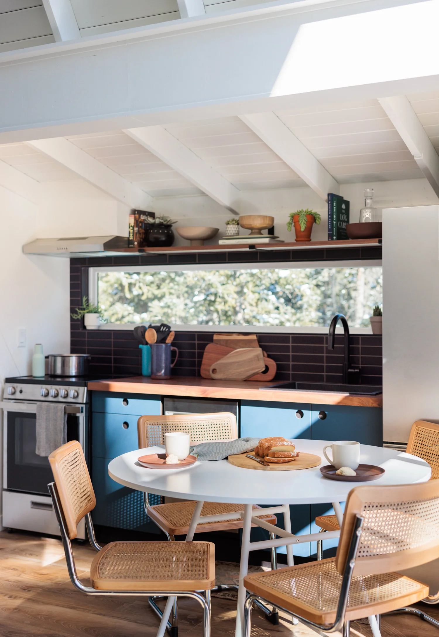 Sitting and dining area with caramel leather seating, round white table, blue cabinets, patterned cushions, and white beams.