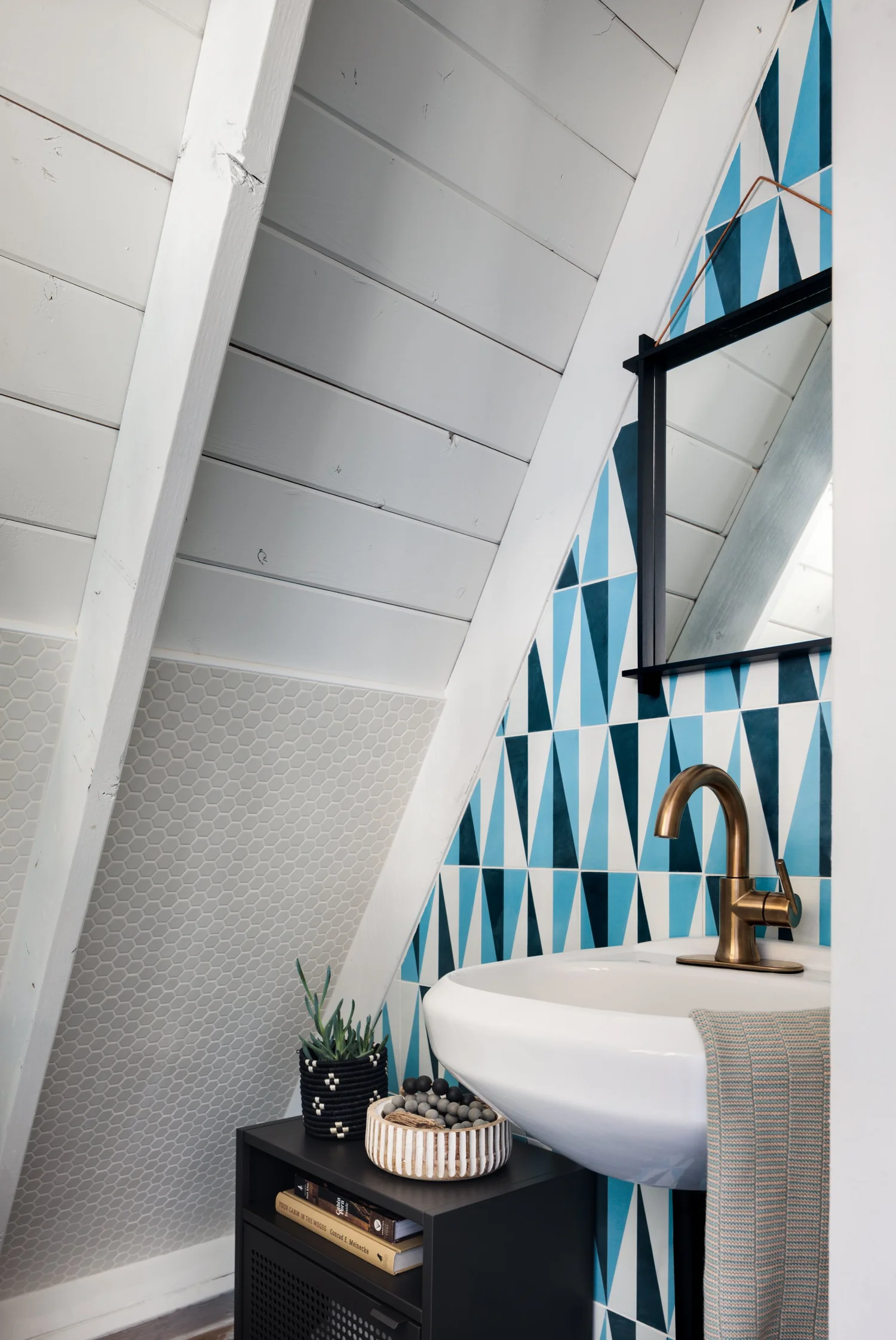 Bathroom with a sloping white wood ceiling, blue geometric tile, rectangular mirror, white sink, and brass faucet.