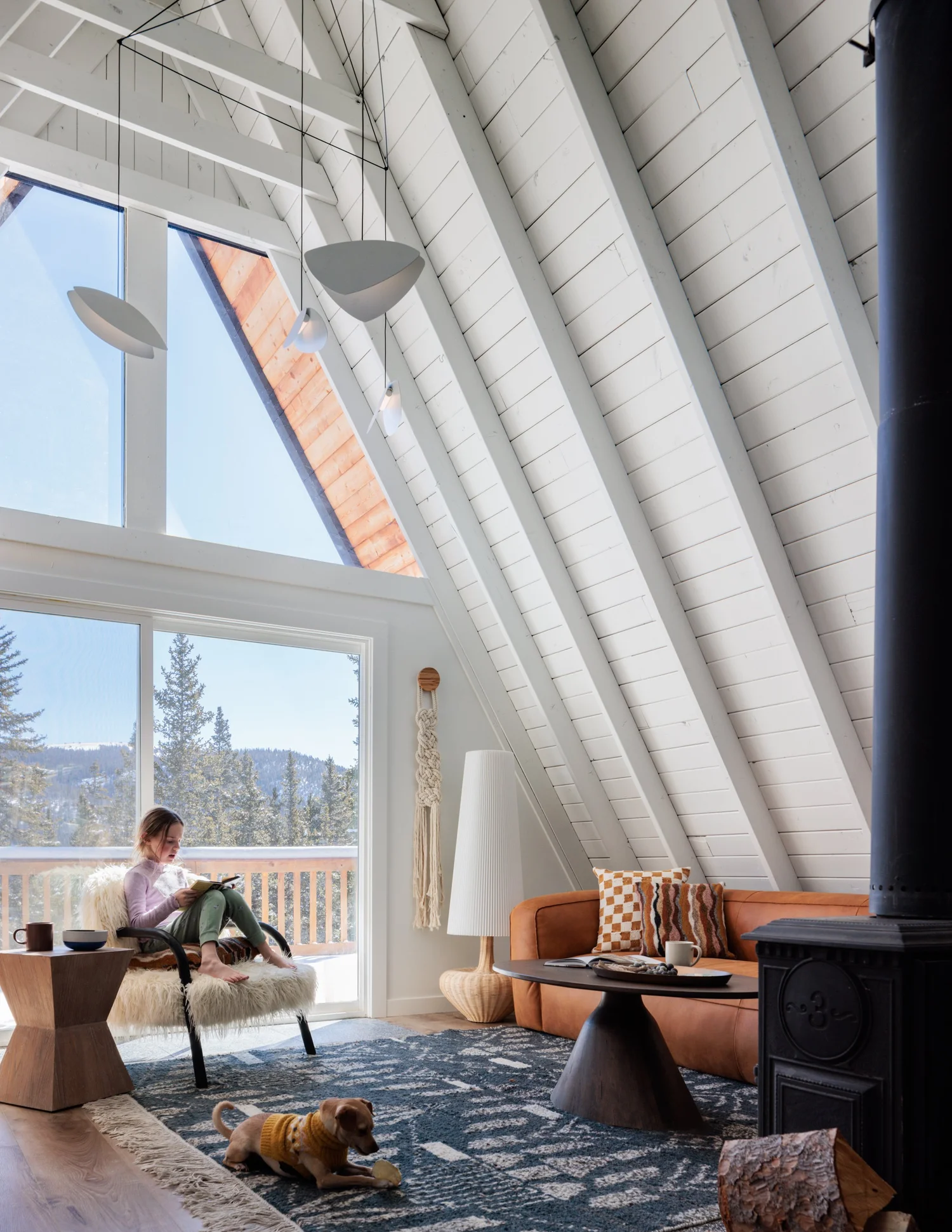 A-frame living room with white ceiling beams, triangular windows, caramel leather seating, black stove, and patterned rug.
