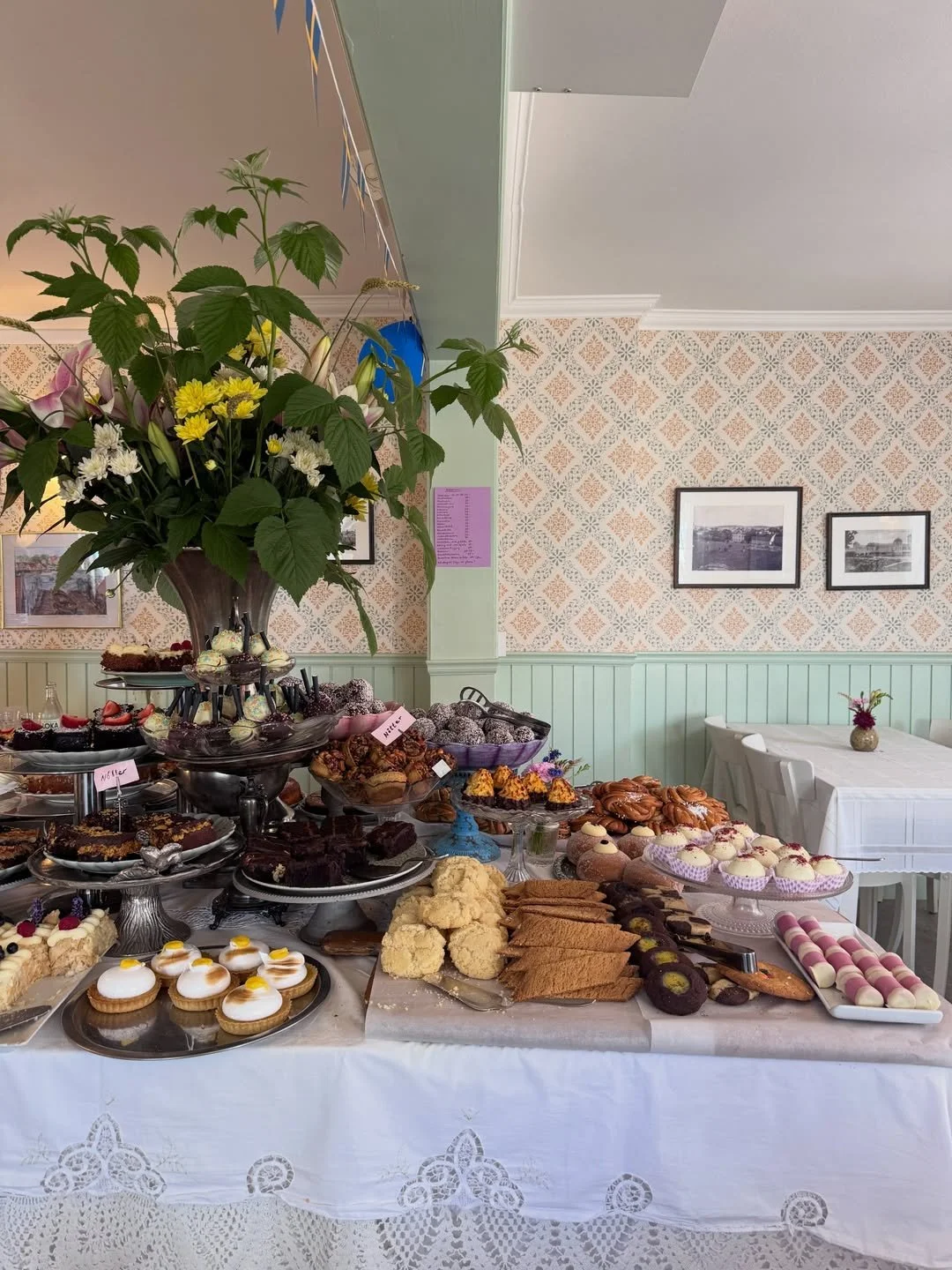 Swedish fika table with pastries, white lace cloth, fresh flowers, patterned wallpaper, and pale green wall paneling.