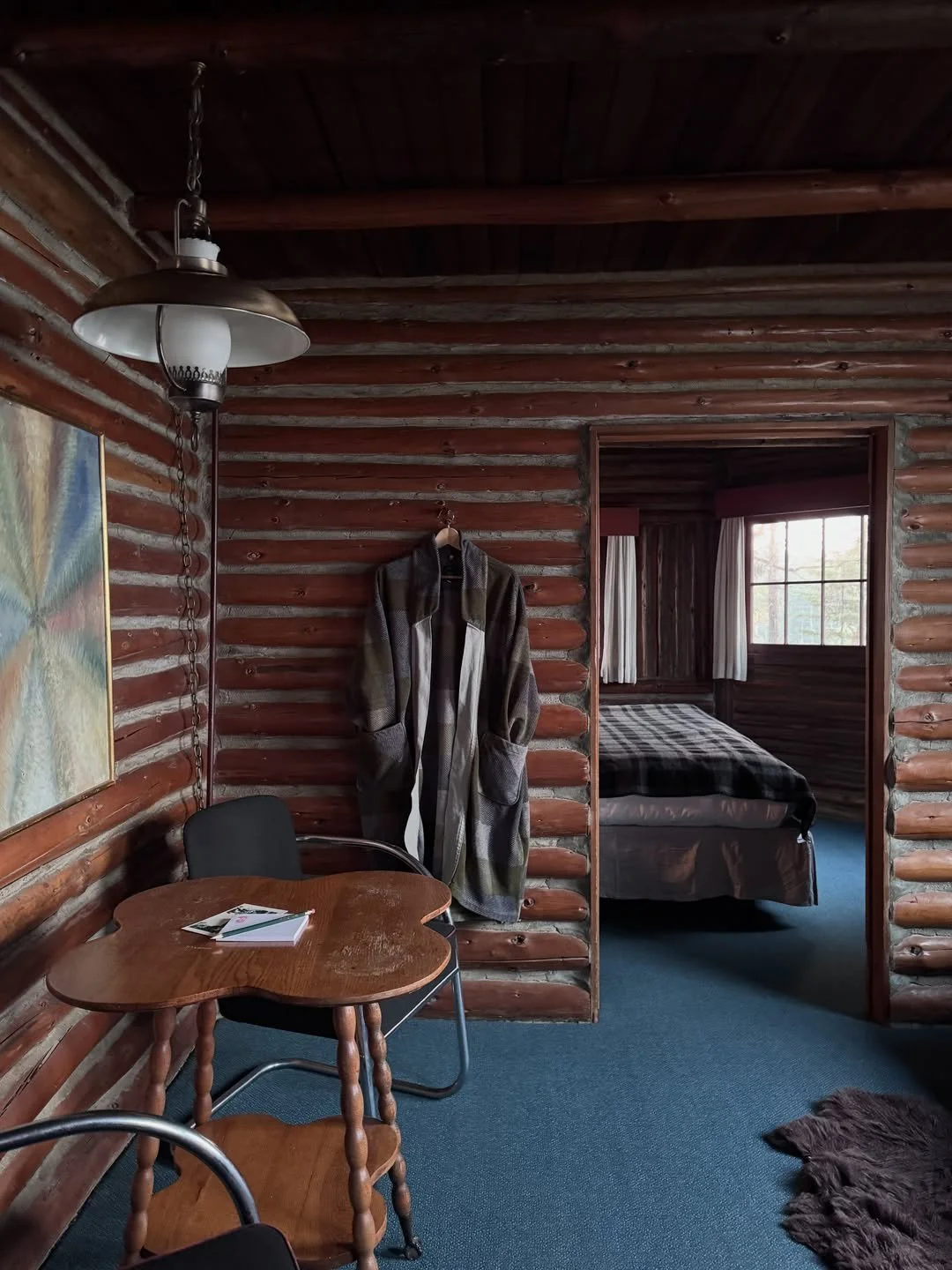 Captain Whidbey log-cabin bedroom with blue carpet, wooden table and chairs, white globe pendant, and dark timber ceiling.