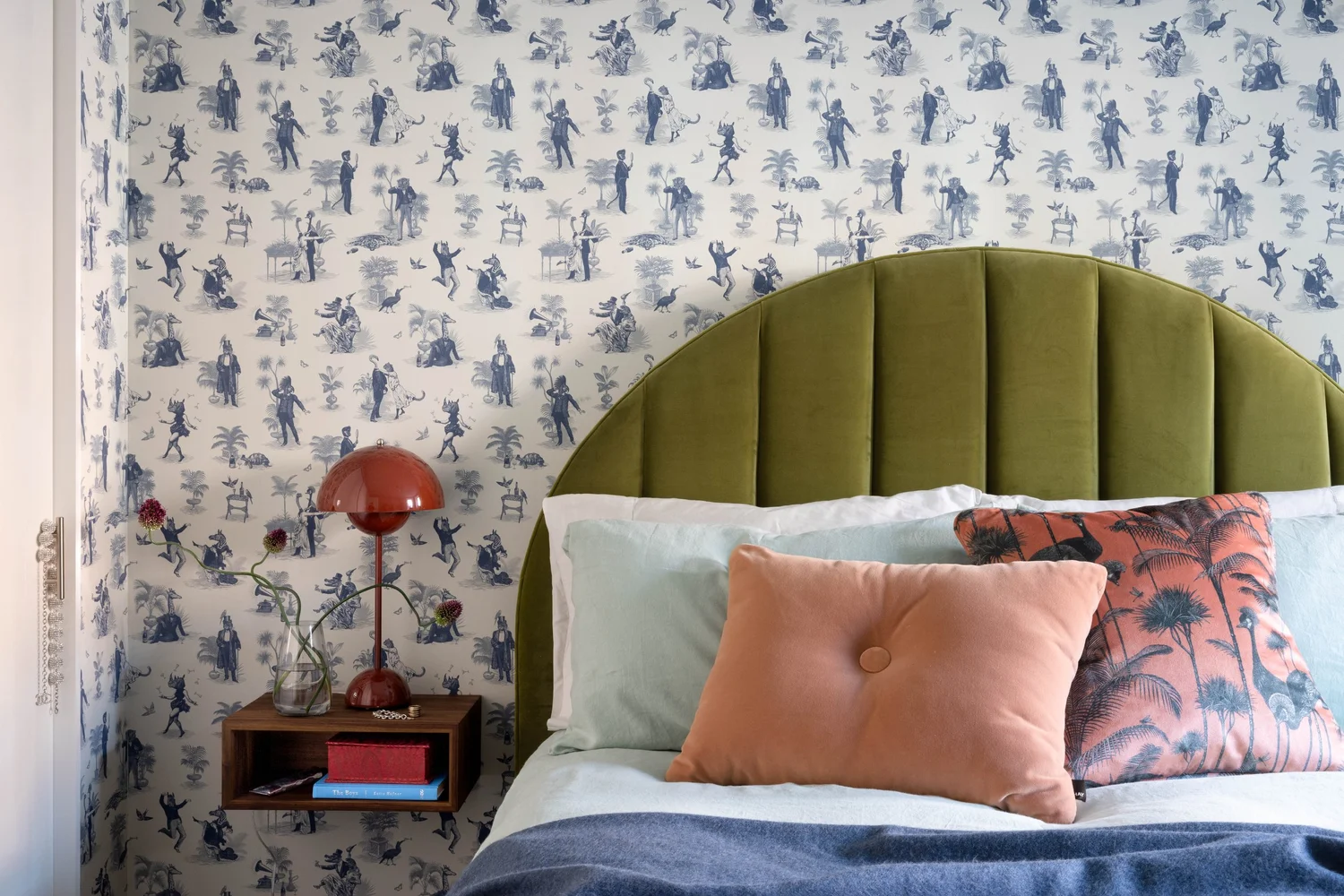 Bedroom with blue-and-white toile wallpaper, green upholstered headboard, pink pillows, blue bedding, and red bedside lamp.