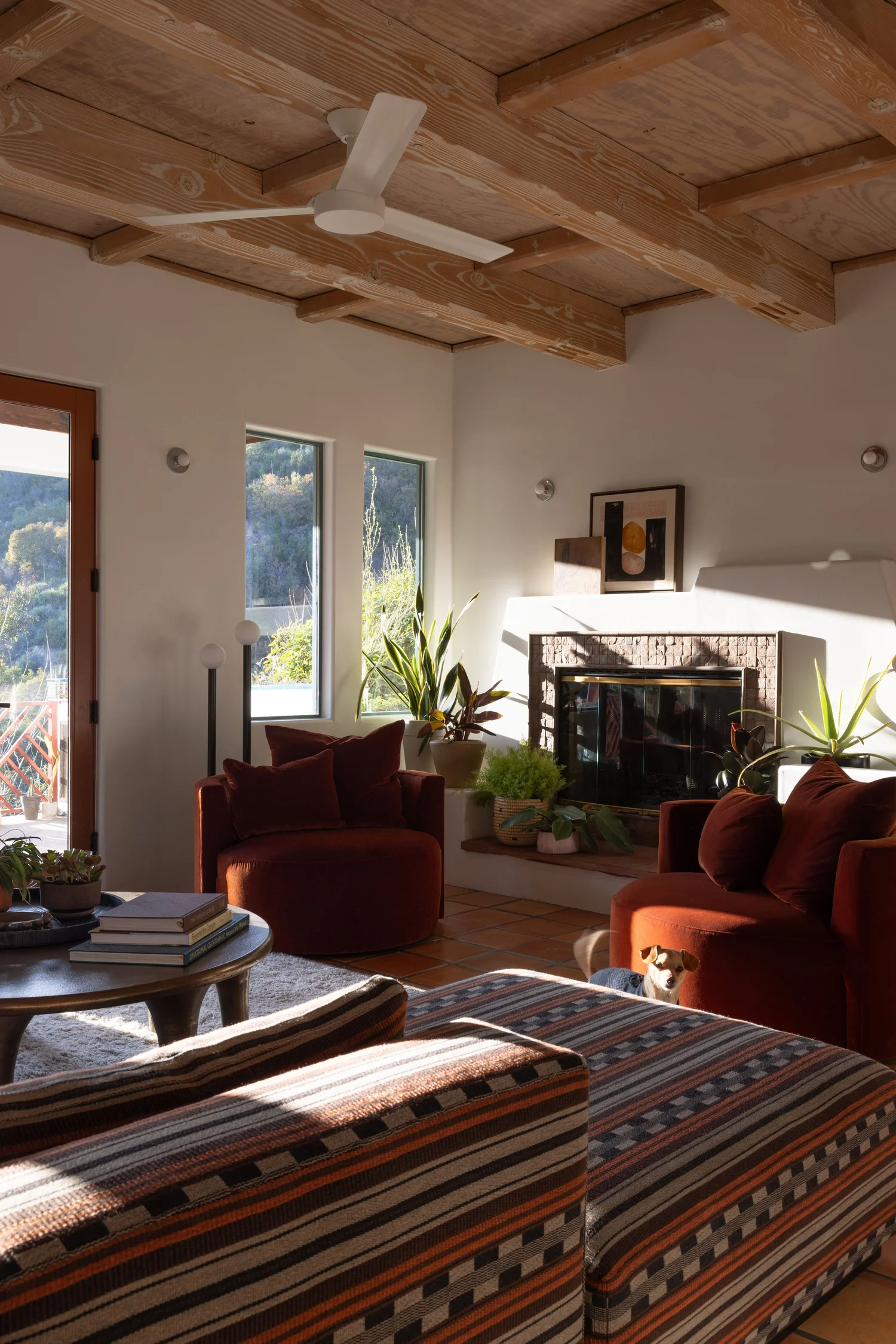 Topanga living room with exposed wood beams, burgundy velvet seating, a corner fireplace, plants, and terracotta flooring.