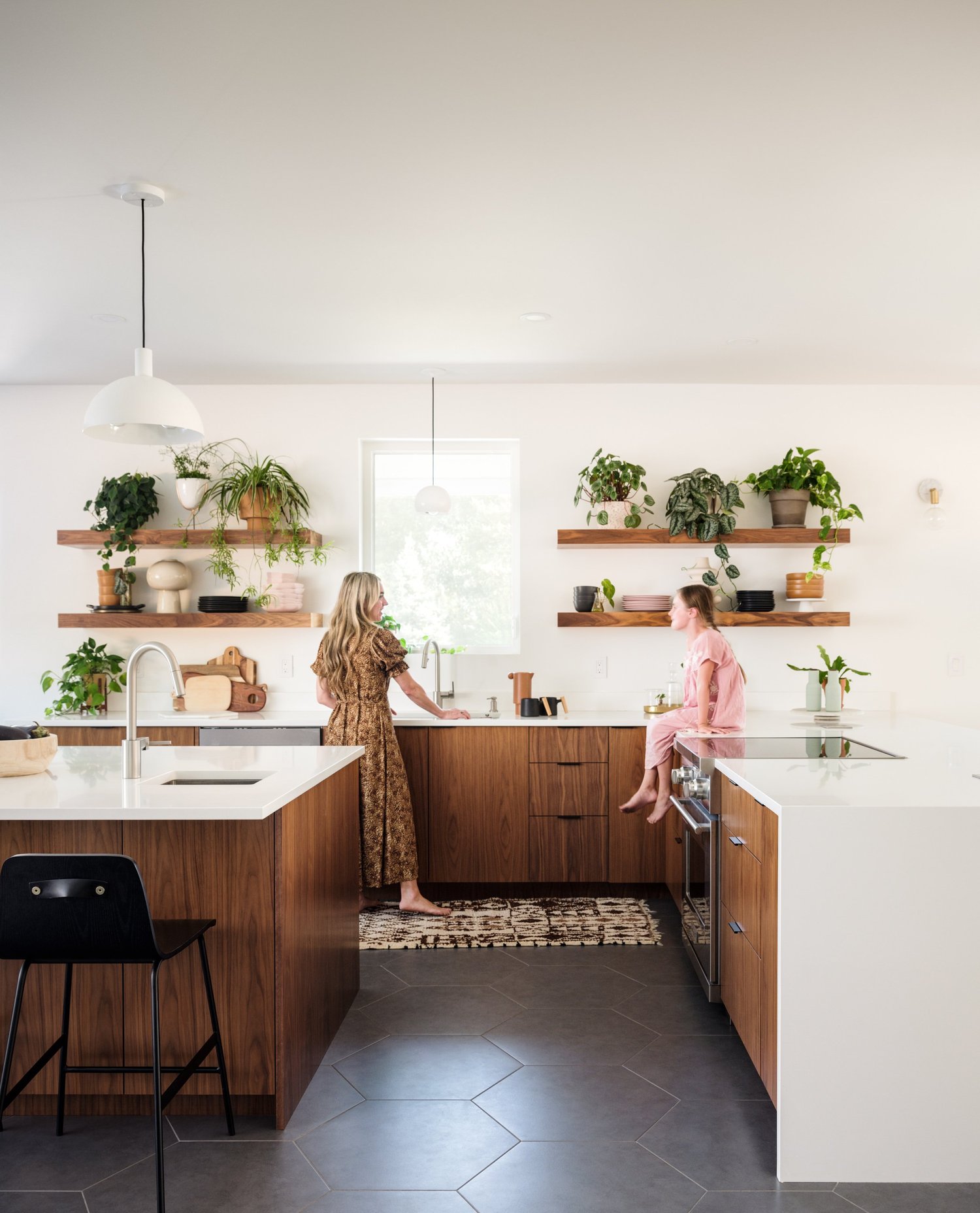 Kitchen with white cabinets, pale counters, open wood shelves, plants, and patterned upholstered dining chairs.