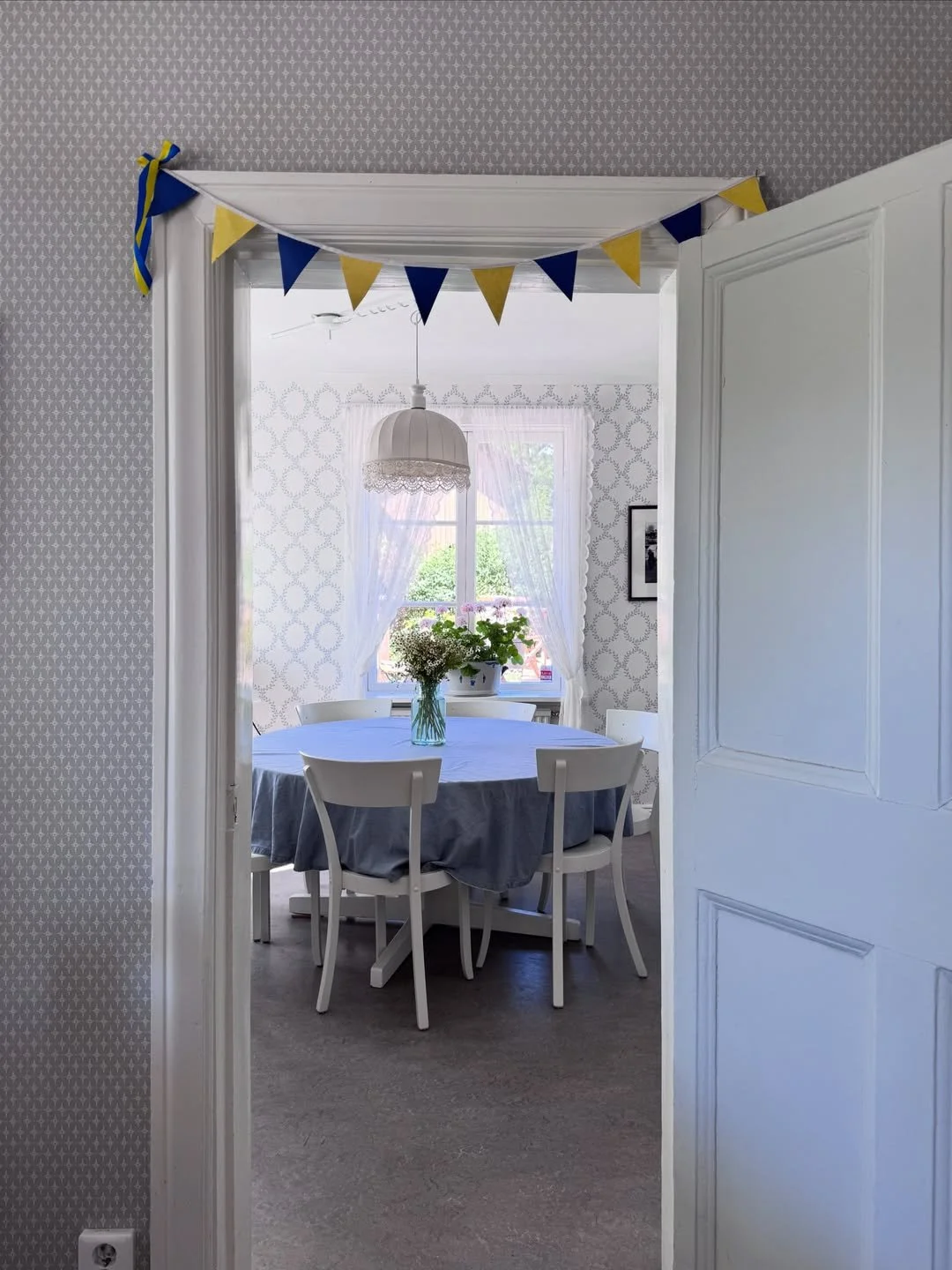 Swedish dining room with patterned wallpaper, white chairs, blue tablecloth, lace pendant shade, and flowers.