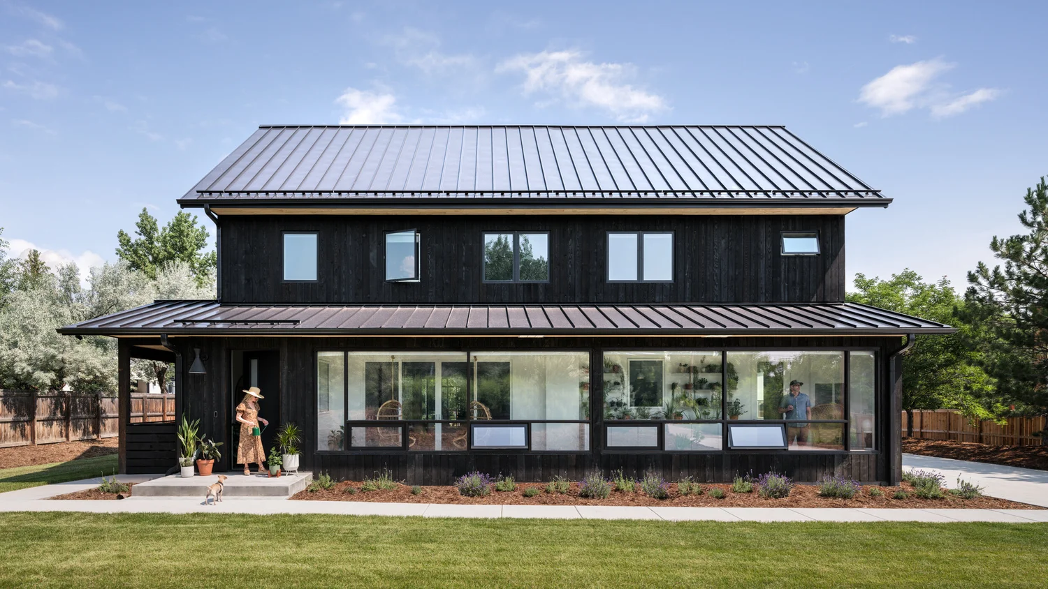 Two-story house with dark siding, metal roof, rows of windows, and covered patio facing the lawn.