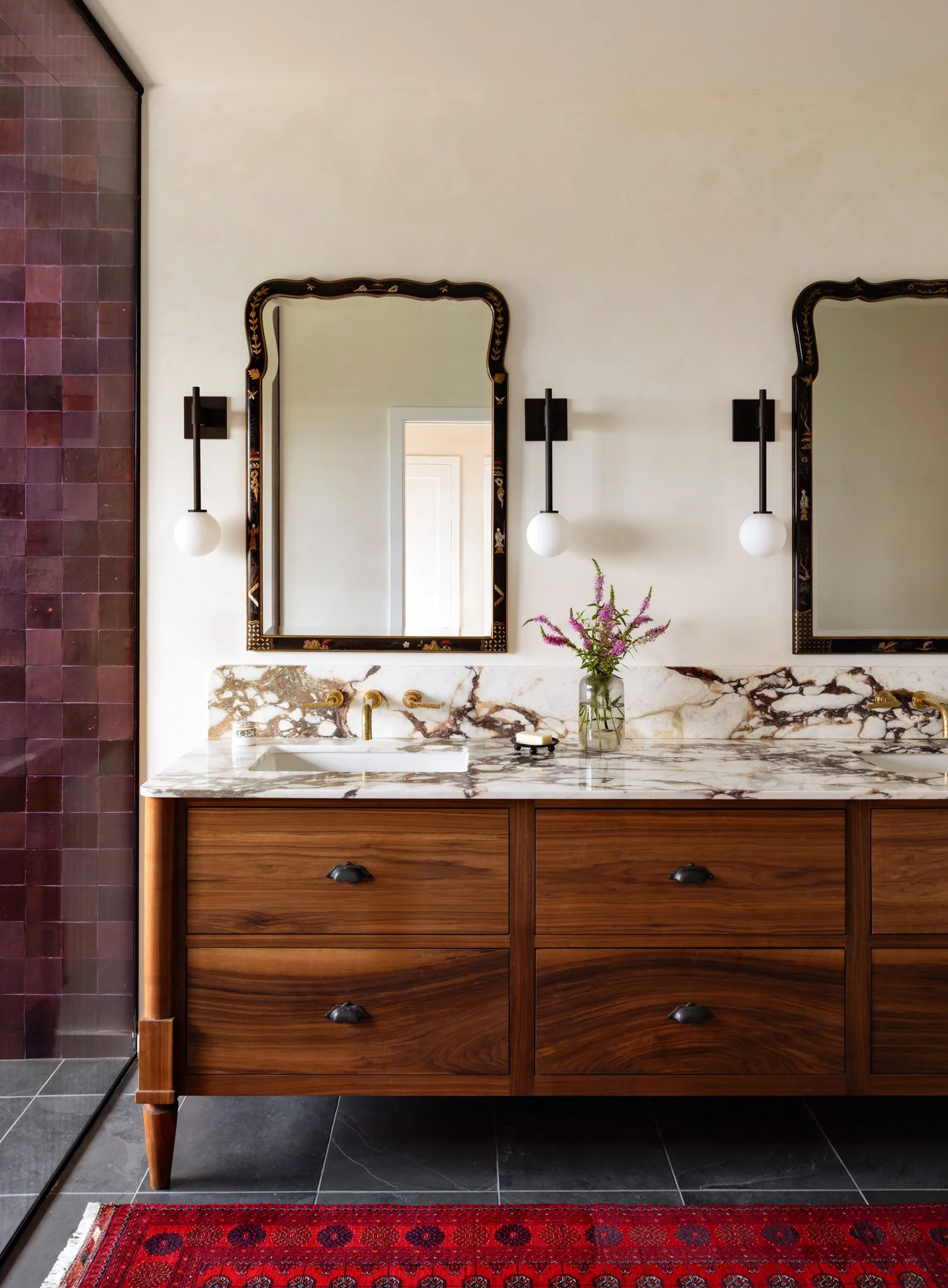 Wood double vanity with veined marble counters, brass faucets, shaped mirrors, and globe sconces.