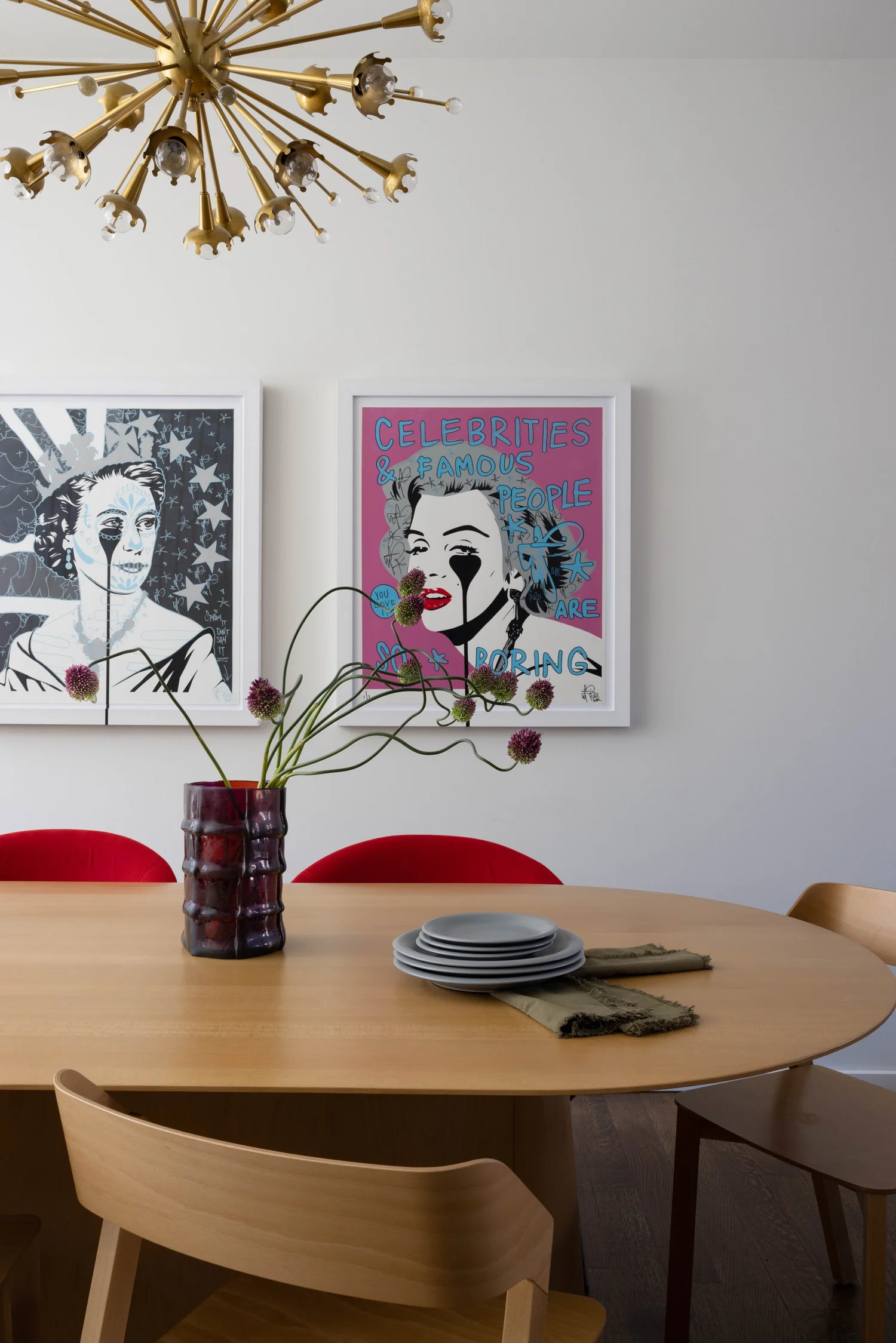 Dining area with a round wood table, curved wood chairs, brass starburst chandelier, pop-art portraits, and flowers.