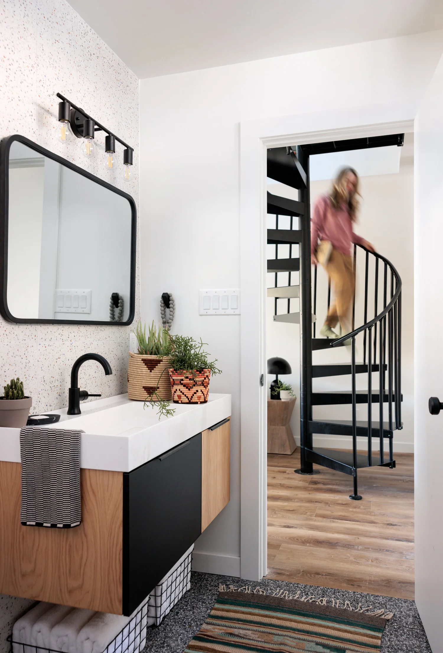 Bathroom with dark vanity cabinet, white basin, rectangular mirror, patterned floor tile, and spiral staircase beyond.