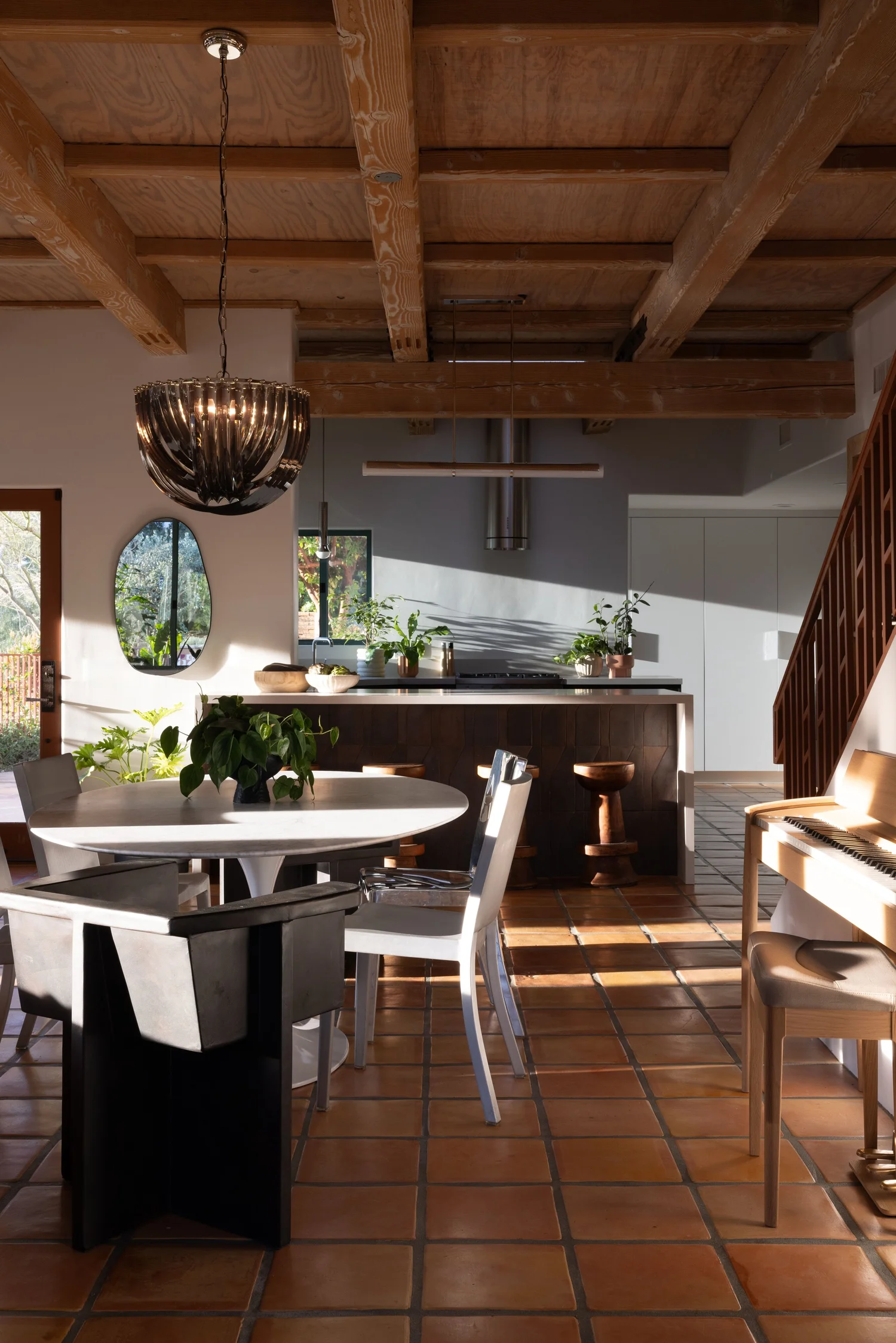 Open kitchen and dining room with exposed wood beams, terracotta floors, a white pedestal table, and sculptural pendant.