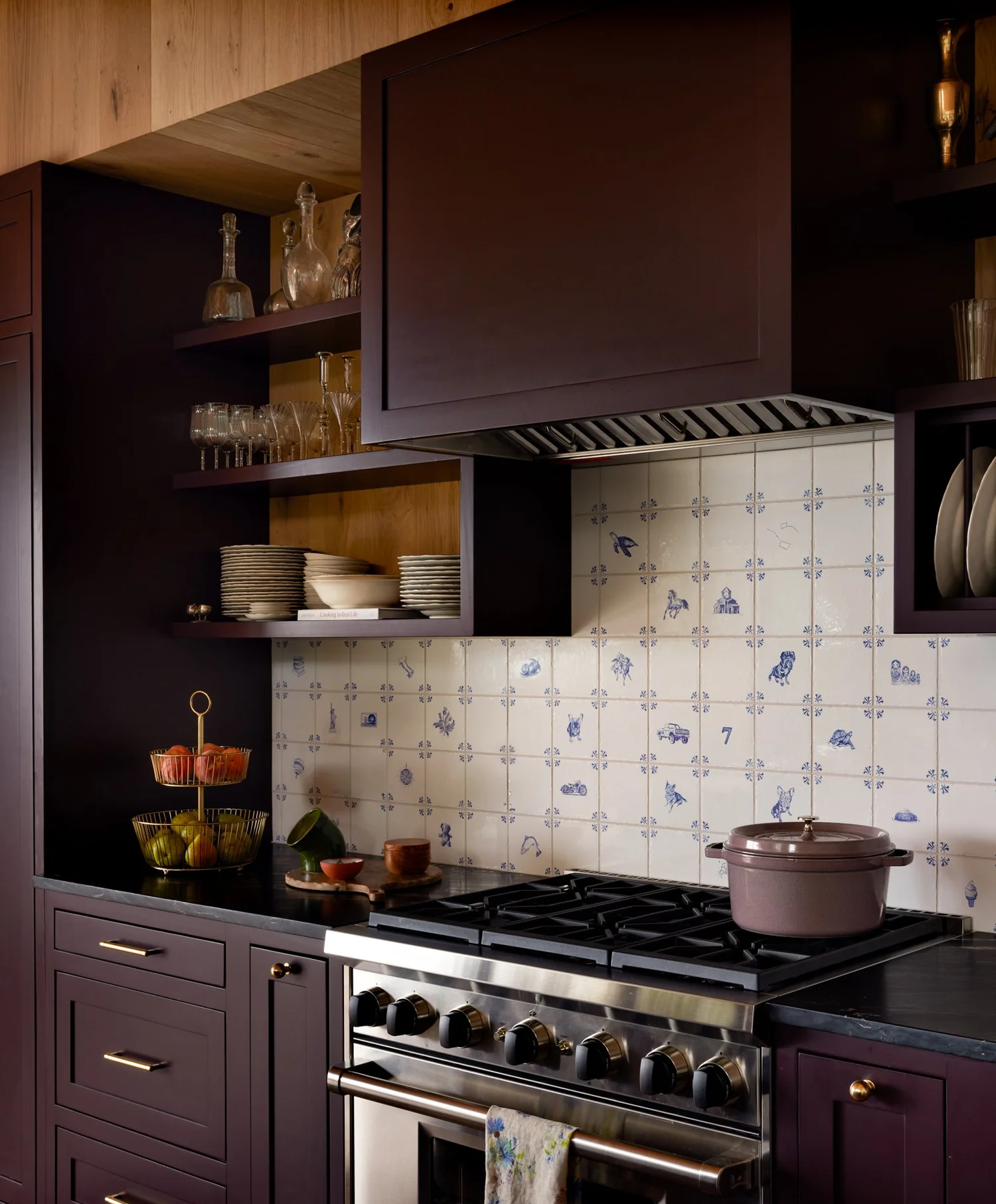 Plum cabinetry, brass hardware, and blue-and-white patterned backsplash beside a stainless steel range.