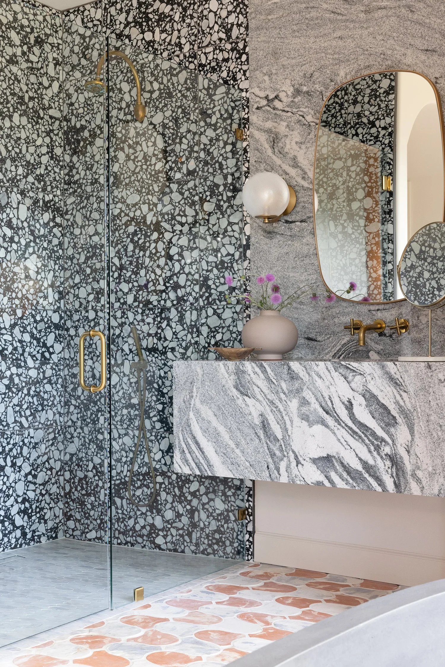 Black-and-white terrazzo shower, veined marble vanity, pink scalloped floor tile, brass fixtures, and irregular mirror.