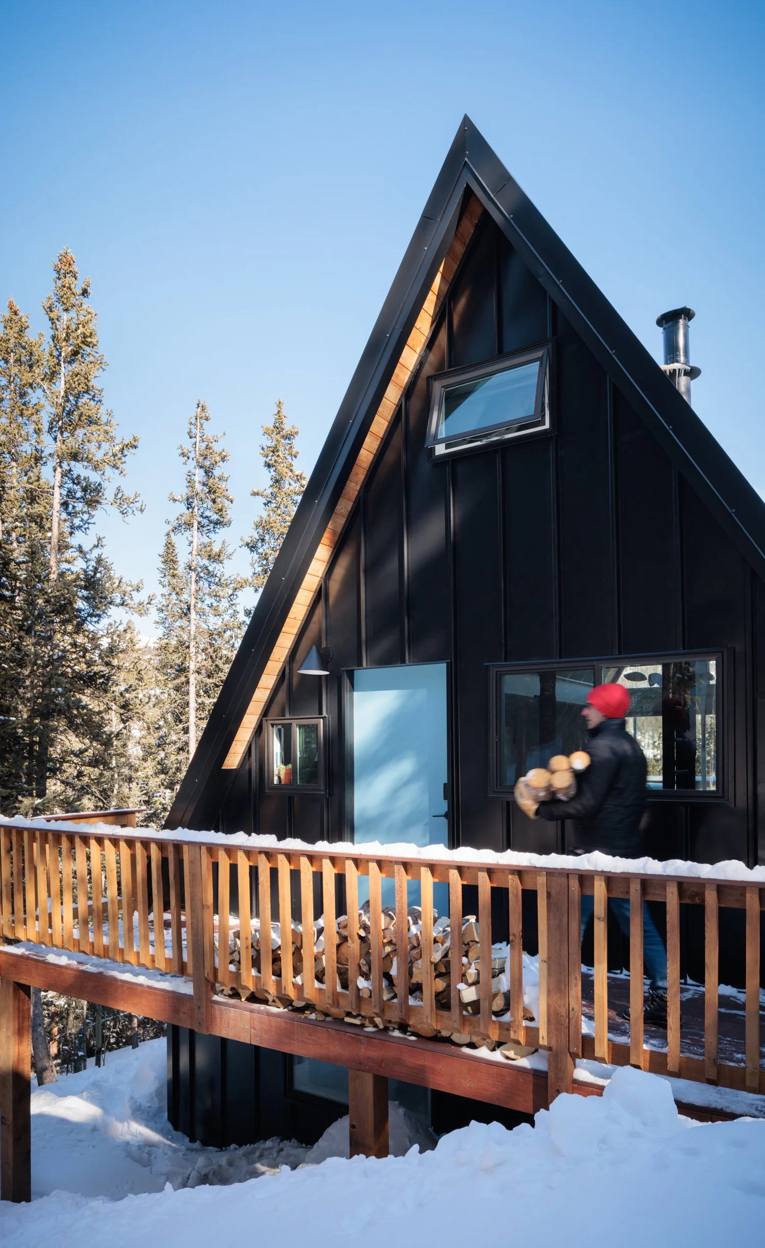 A-frame cabin with black siding, triangular windows, wood deck railing, and surrounding pine trees.