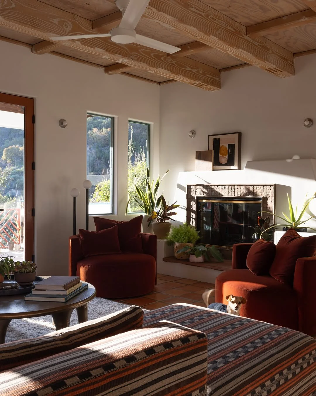 Living room with Danielle Hutchens art above a brick fireplace, burgundy velvet chairs, patterned upholstery, plants, and wood beams.