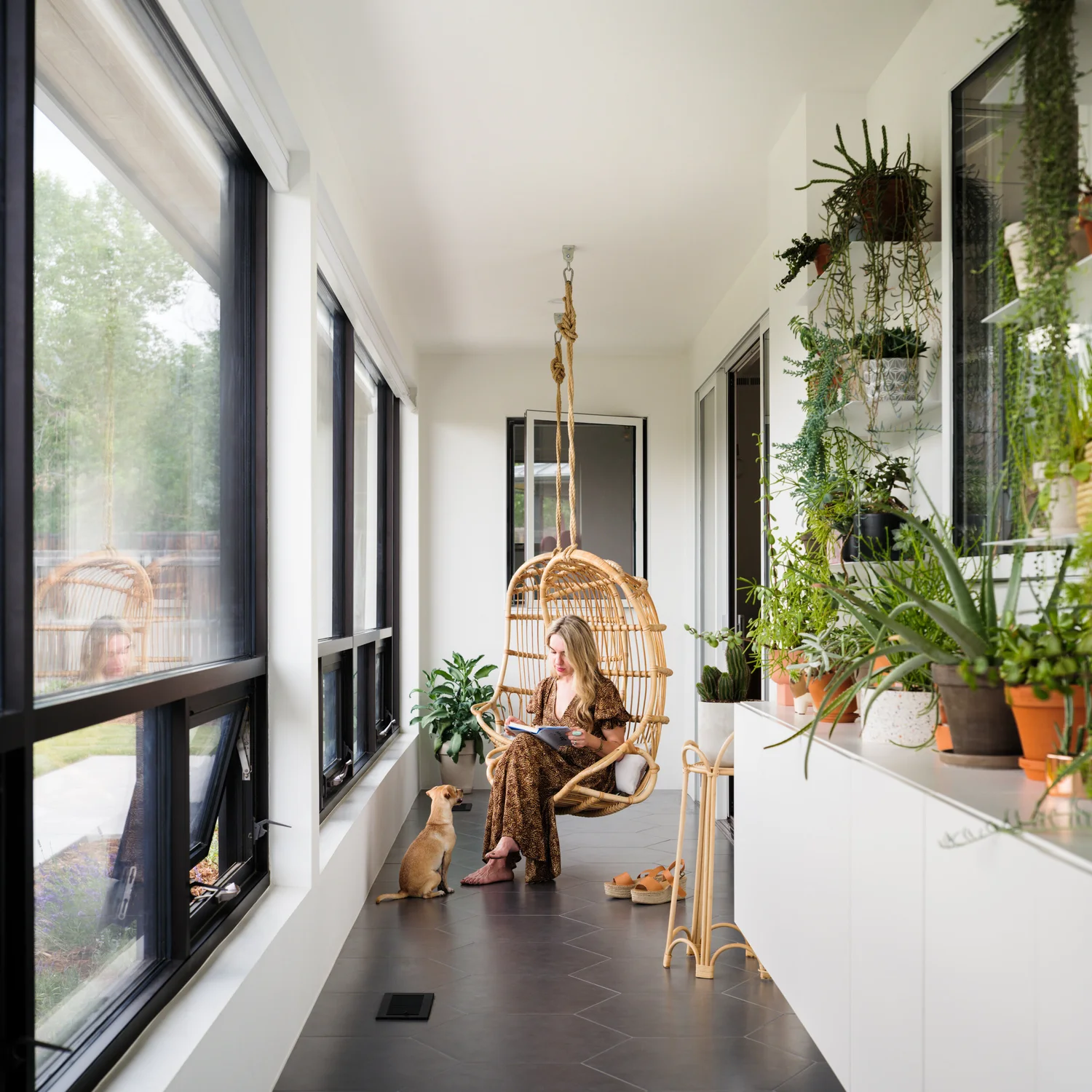 Enclosed porch with tall black-framed windows, pale flooring, wood furnishings, and clusters of potted plants.