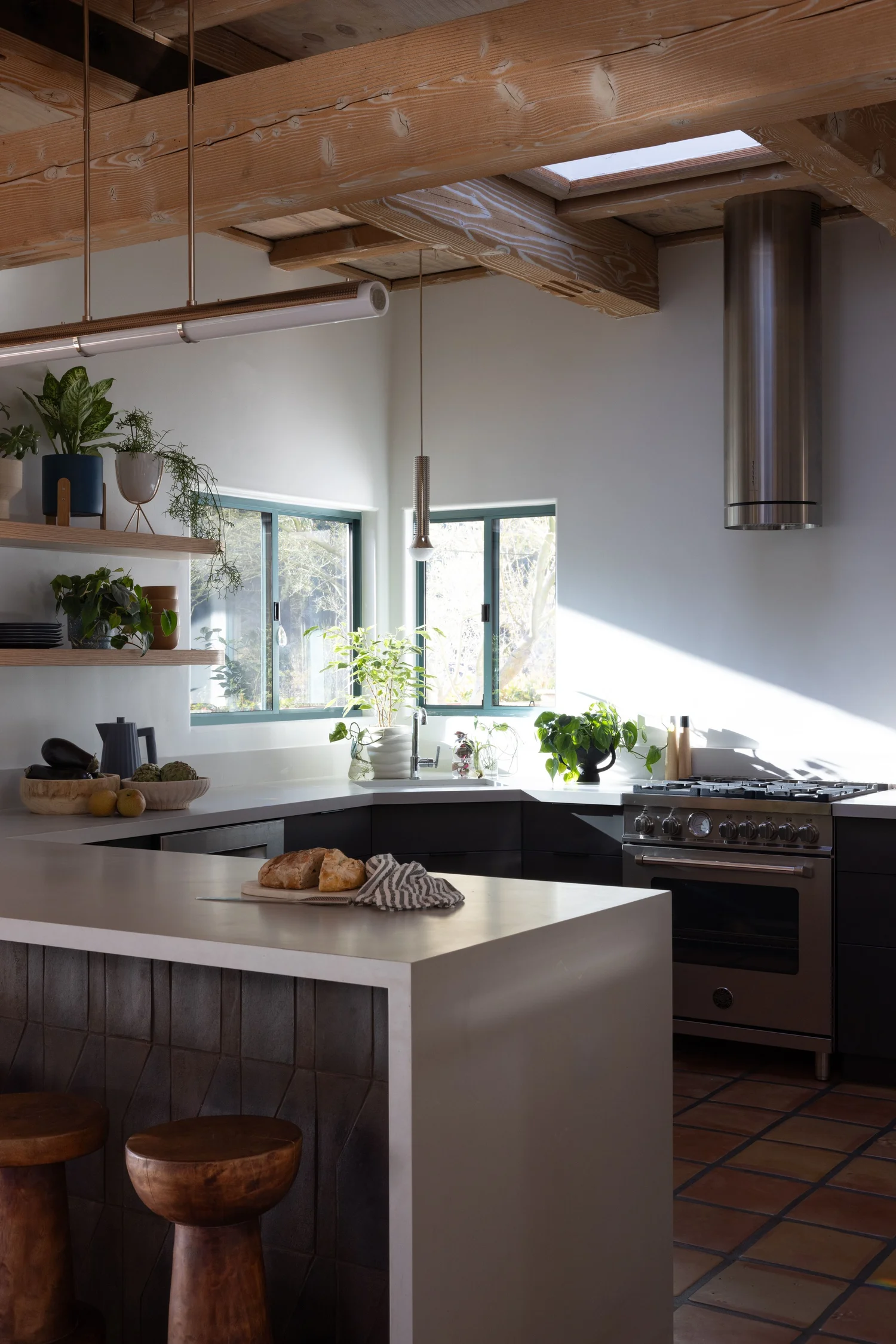 Kitchen with exposed wood beams, a white waterfall island, wood stools, dark cabinets, and open shelving.