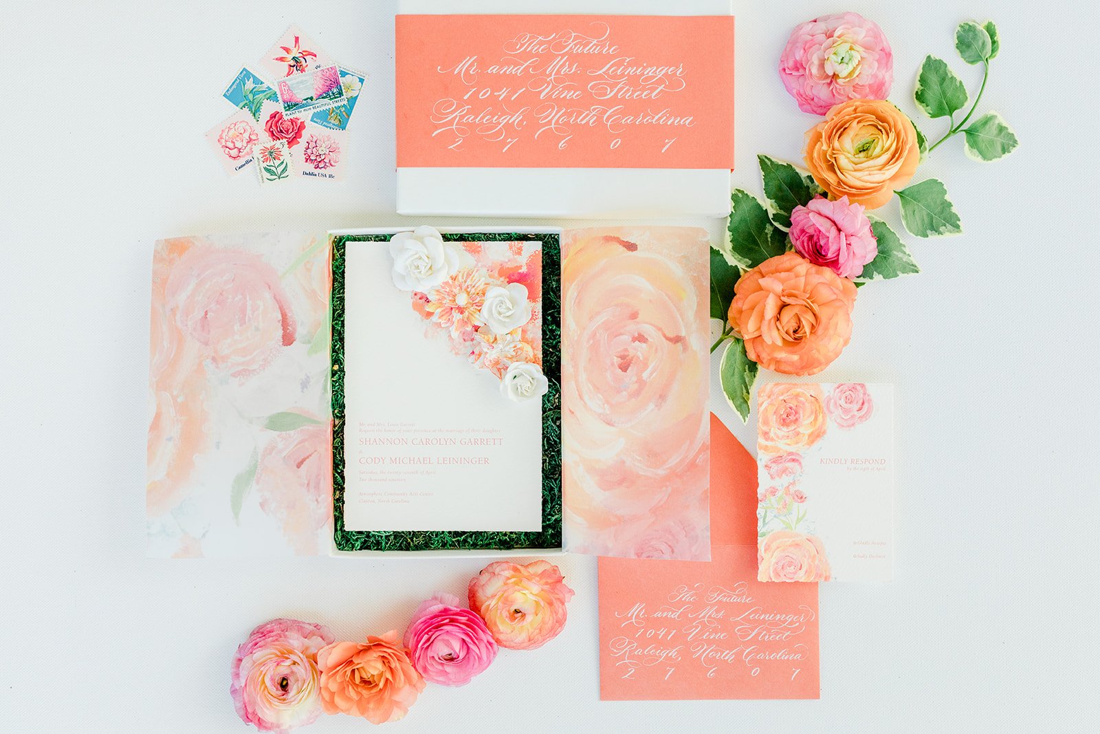 A flat lay of a wedding invitation suite with bright orange envelopes and calligraphy. The main invitation has 3D paper flowers and pink and orange painted textures. The typography is clean serif type in a deep pink.