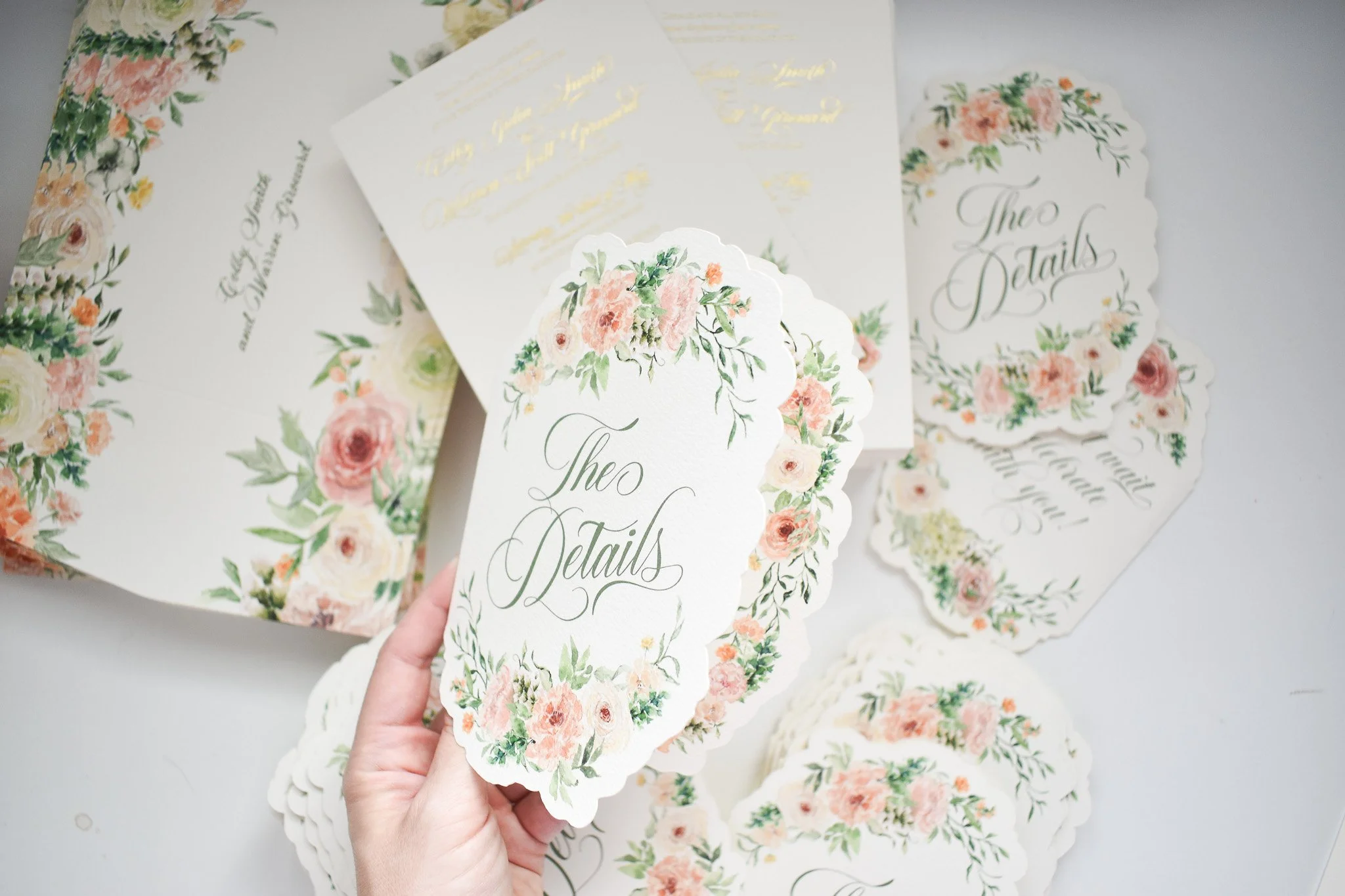A hand holds a die-cut, scalloped wedding details card featuring a watercolor border of blush roses and greenery with elegant green script calligraphy. In the background are coordinating gold foil invitations and patterned envelopes.
