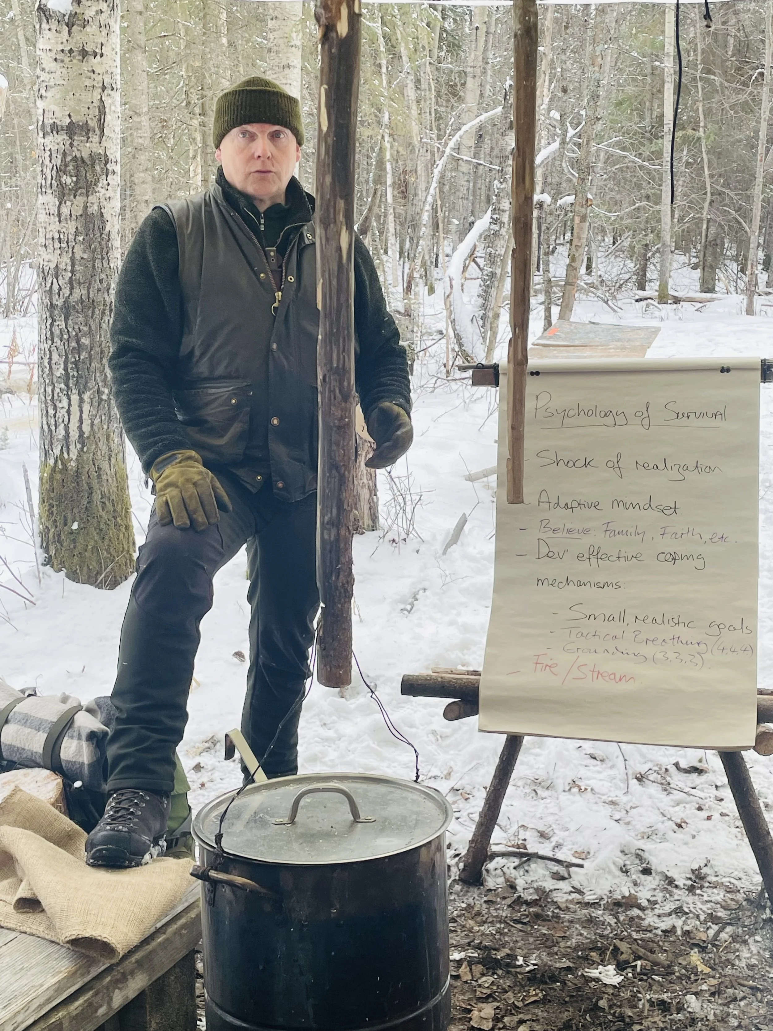 Man dressed warmly in a beanie, gloves, and black vest stands outdoors in a snowy forest next to a campfire pot and a wooden board with notes. The notes discuss psychology of survival, adaptive mindset, achievable goals, and fire/stream.