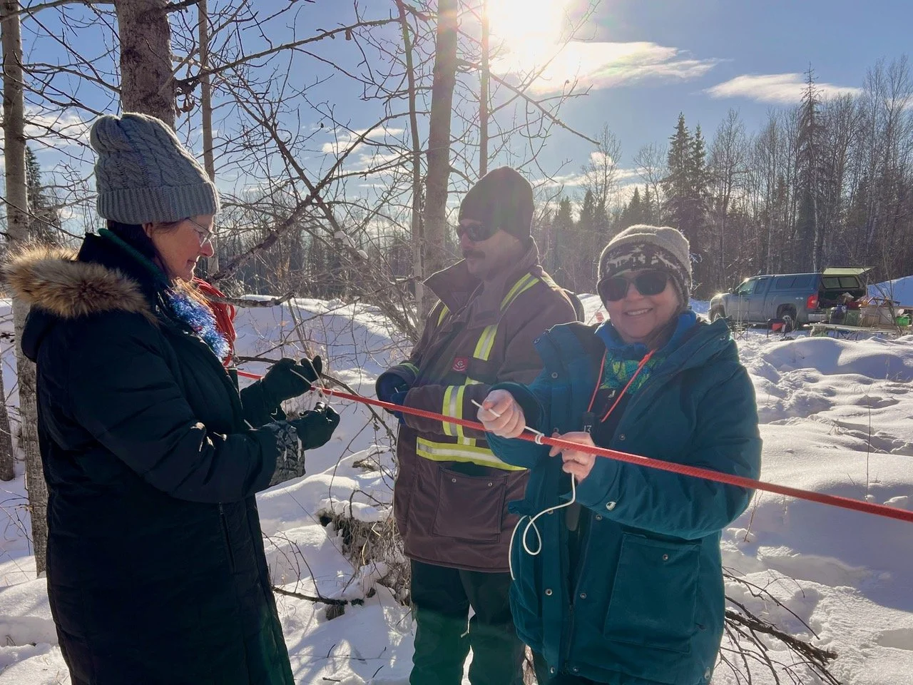 Three people outdoors in winter, dressed in warm clothing, standing among snow-covered trees, holding a red rope, learning knots with vehicles and equipment in the background.