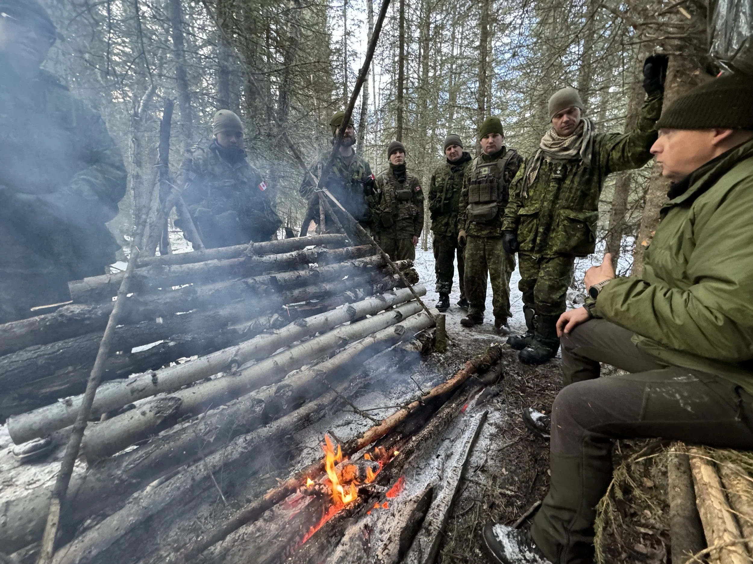Group of eight soldiers in camouflage military uniforms with some wearing Canadian flags, gathered around a small campfire in a forest during winter, with snow on the ground and leafless trees, under a cloudy sky.