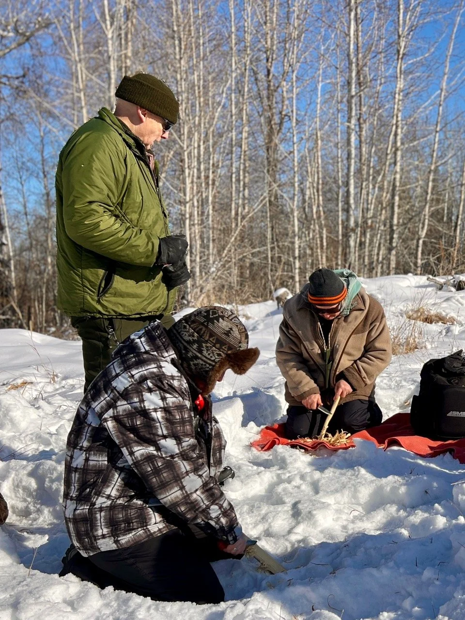 Three people in winter gear are engaged in an outdoor activity in a snowy forest. One person is standing, wearing a green jacket and a knit hat, while two others are kneeling and working with tools on the snow.