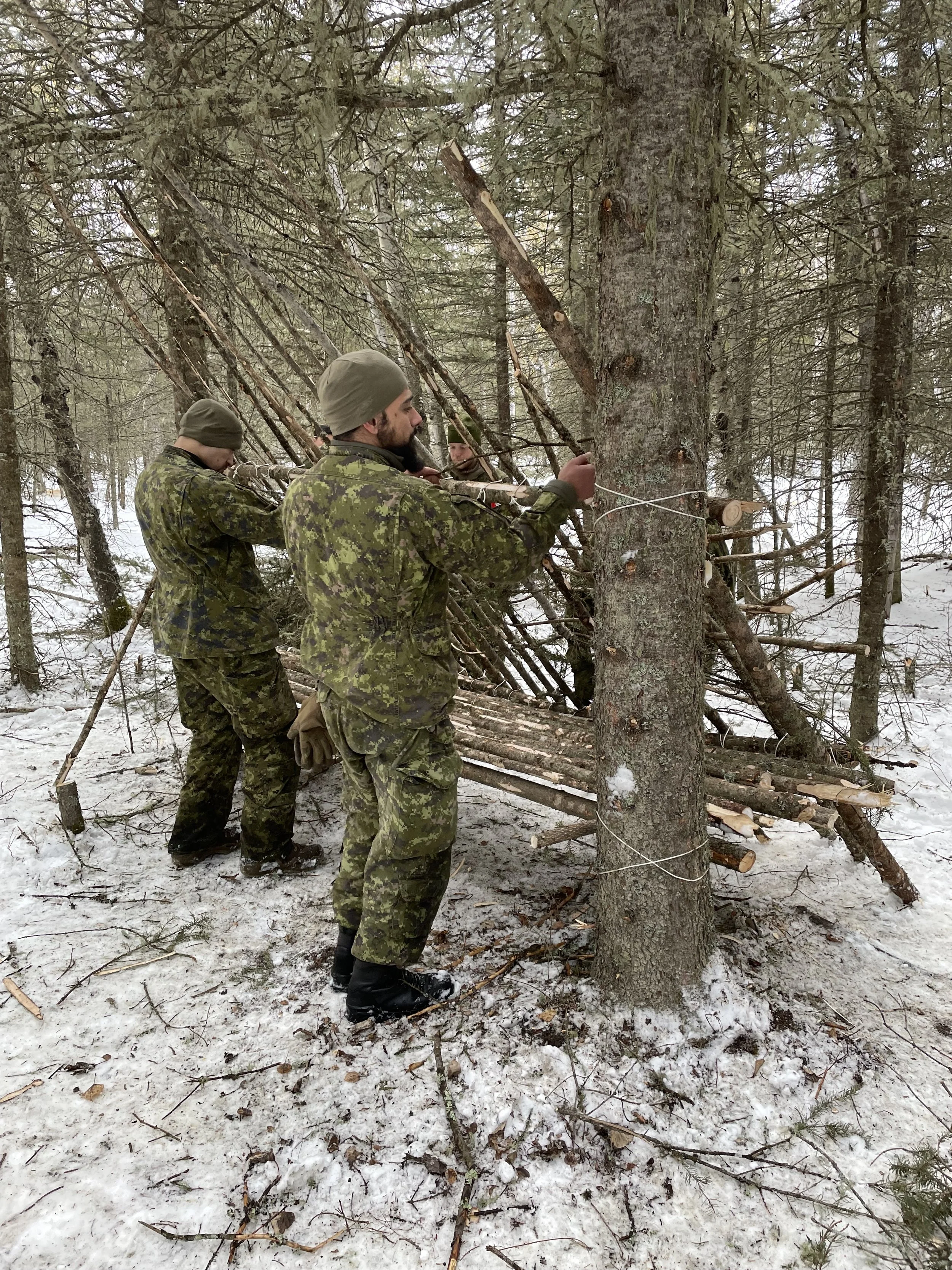 Three soldiers in camouflage uniforms building a camouflaged shelter in a snowy forest, using logs and rope.