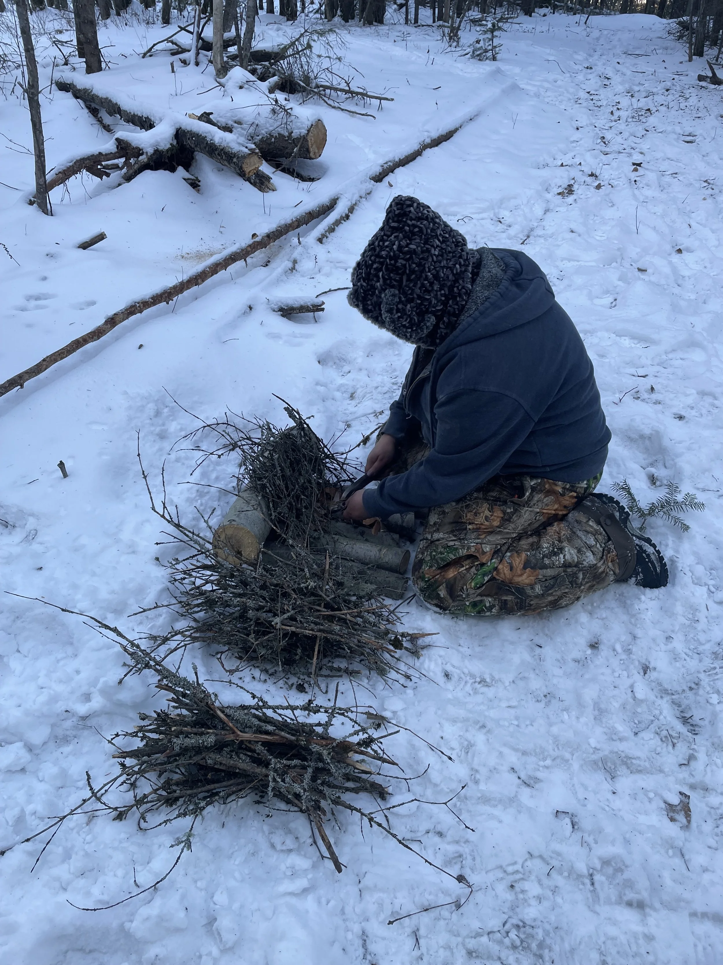 A person kneeling in the snow, gathering sticks and branches with a pile of wood next to them in a wooded area.