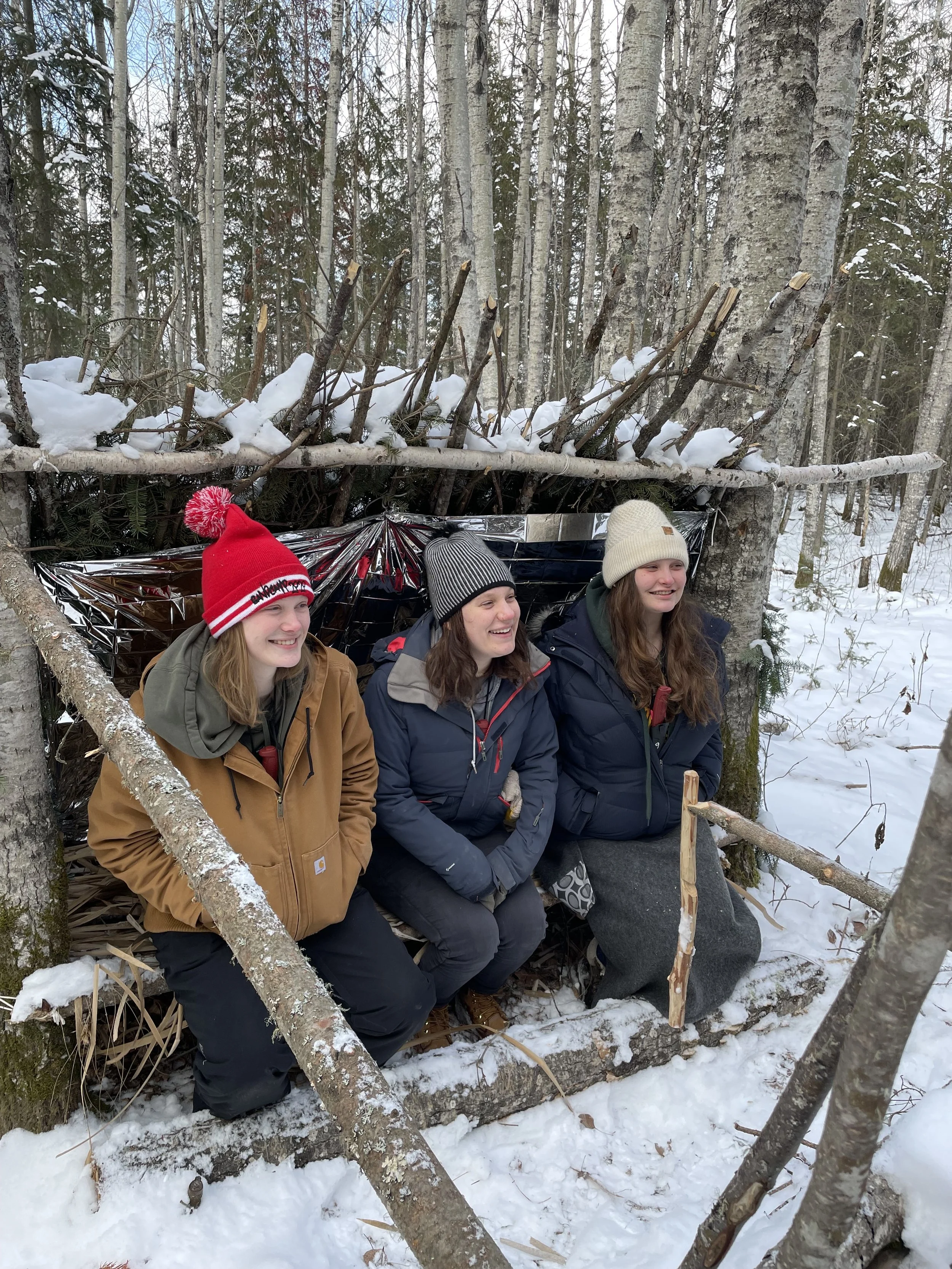 Three young women sitting in a snow-covered forest, smiling, with a lean-to shelter made of sticks and a reflective blanket behind them.