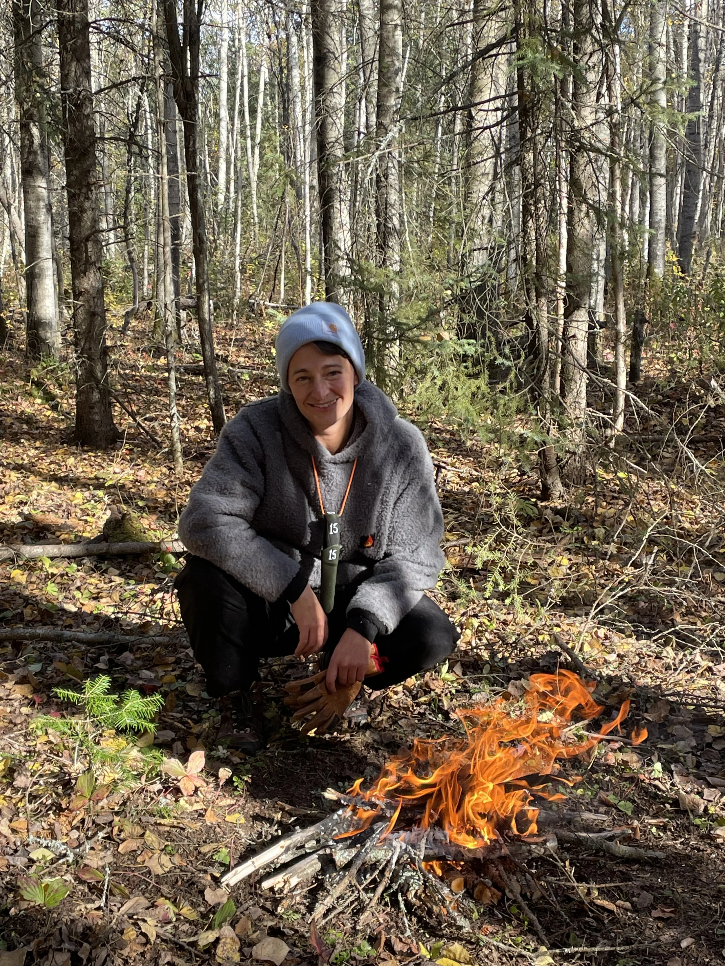 A woman with a gray beanie and jacket squats in a forest next to a small campfire with flames, surrounded by fallen leaves and trees.
