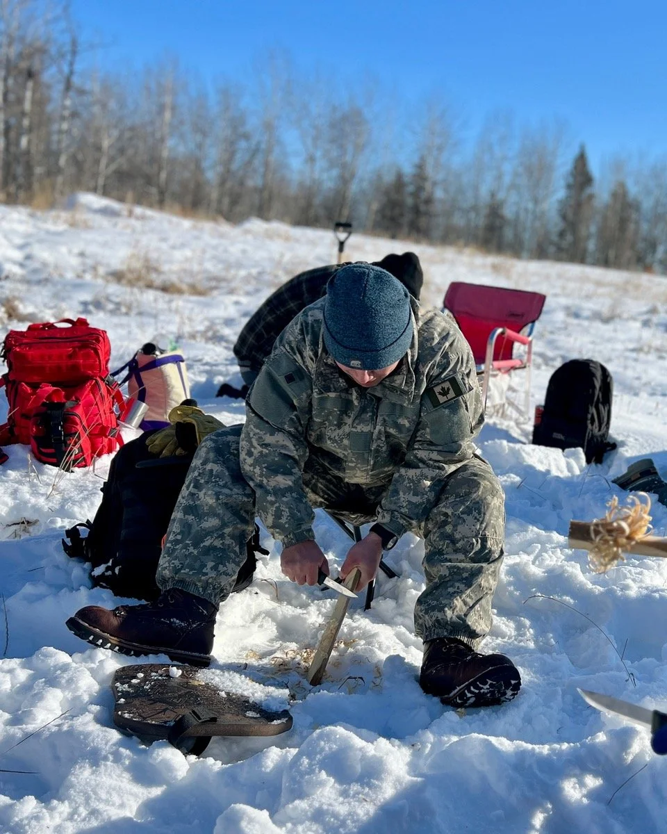 A person in camouflage military uniform and a gray beanie using a knife to cut feather sticks on a cold winter day. There are backpacks, a red chair, and various gear in the background, with a snowy field and leafless trees under a clear blue sky.