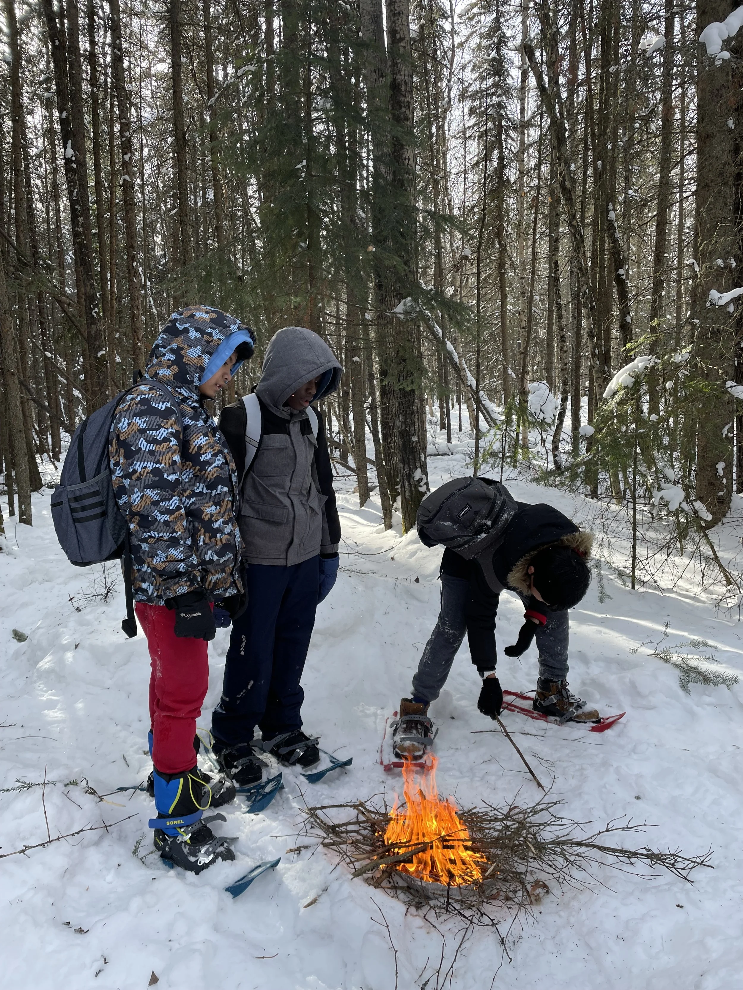 Three hikers in winter clothing standing around a small campfire in a snowy forest, with one tending the fire and two observing.