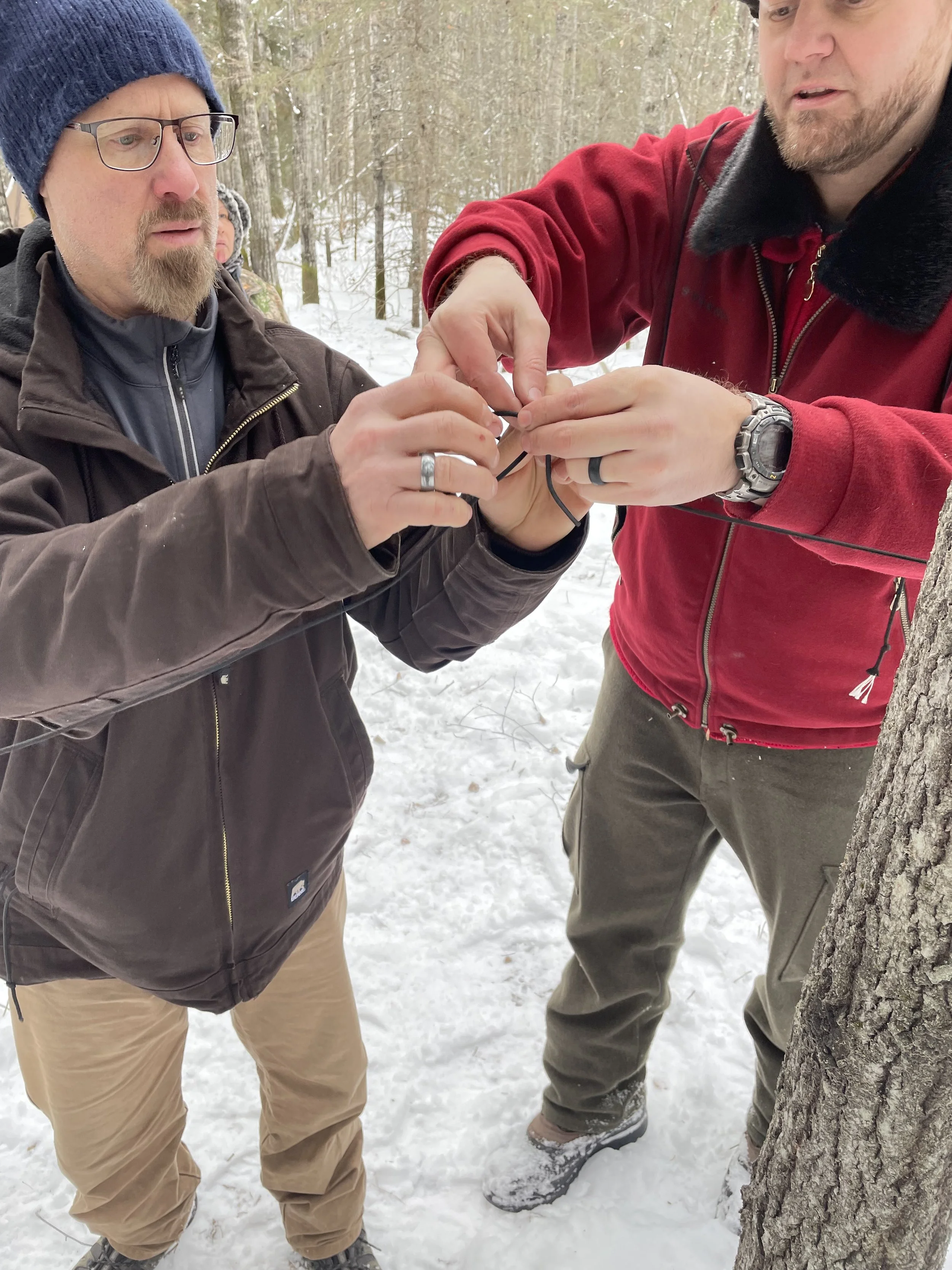 Two men working together outdoors in a snowy forest, handling a small black electronic device or wire.