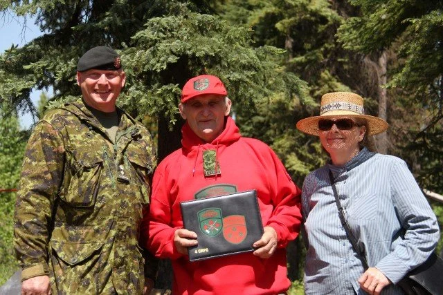 Mors Kochanski and his wife Dianna, smiling at the camera. Mors is dressed in a red Canadian Ranger red hoodie and cap holding a certificate with military patches, and the Dianna is wearing sunglasses, a straw hat, and a light blue shirt.