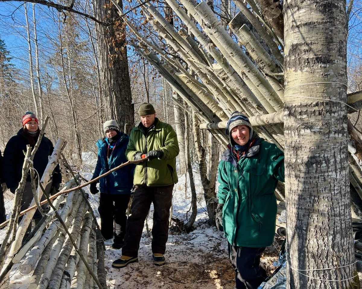 Four people in winter clothing standing outdoors in a snowy forest, constructing a shelter using fallen tree branches and logs.
