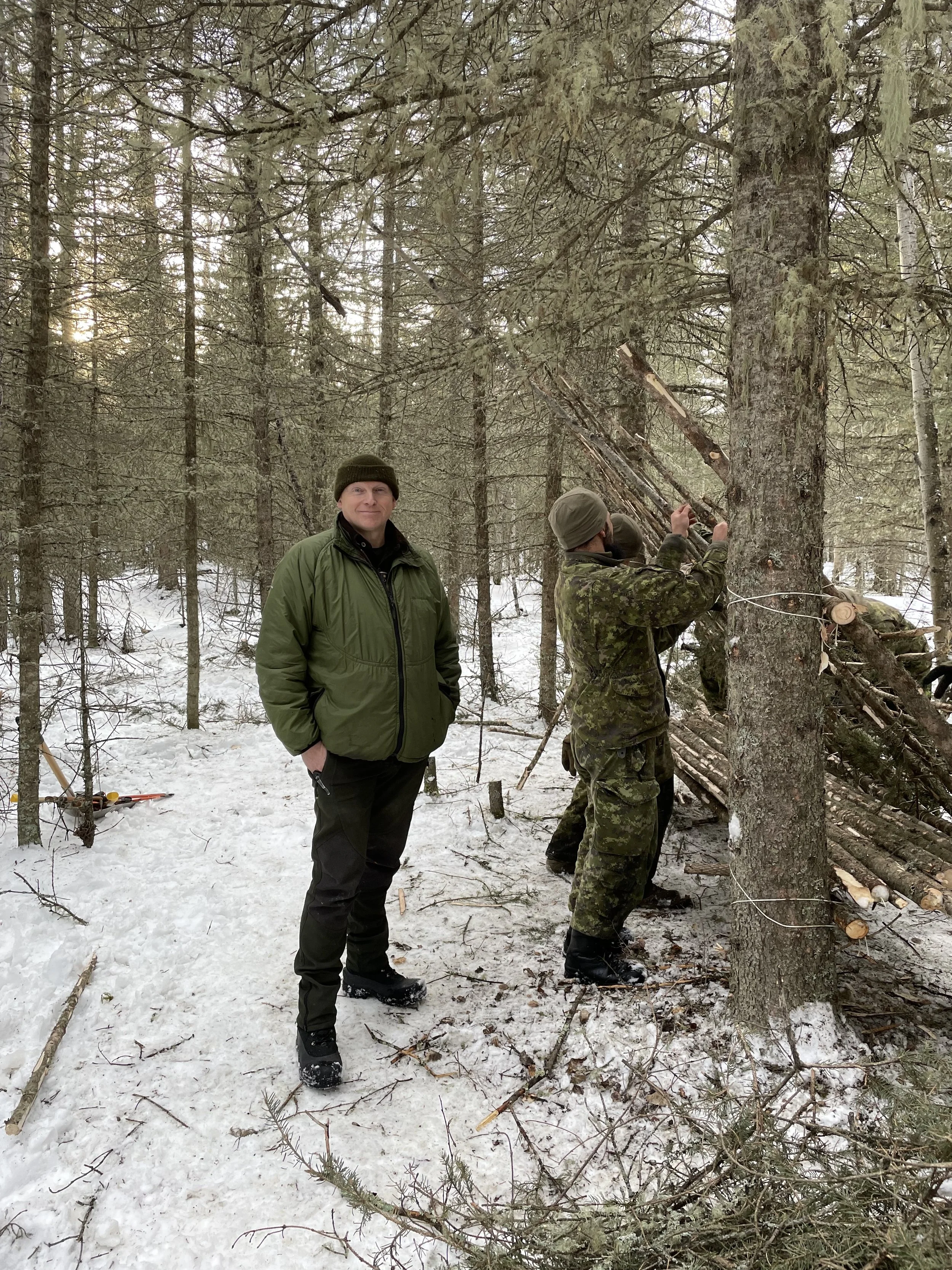 A man in a green jacket and black pants standing in a snowy forest, with another person in camouflage cutting a tree with a saw. There are snow-covered ground and numerous pine trees surrounding them.