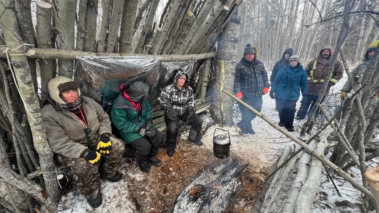 Group of people in winter clothing sitting and standing around a makeshift shelter made of sticks and plastic, with a small fire burning on the snowy ground in a forest.